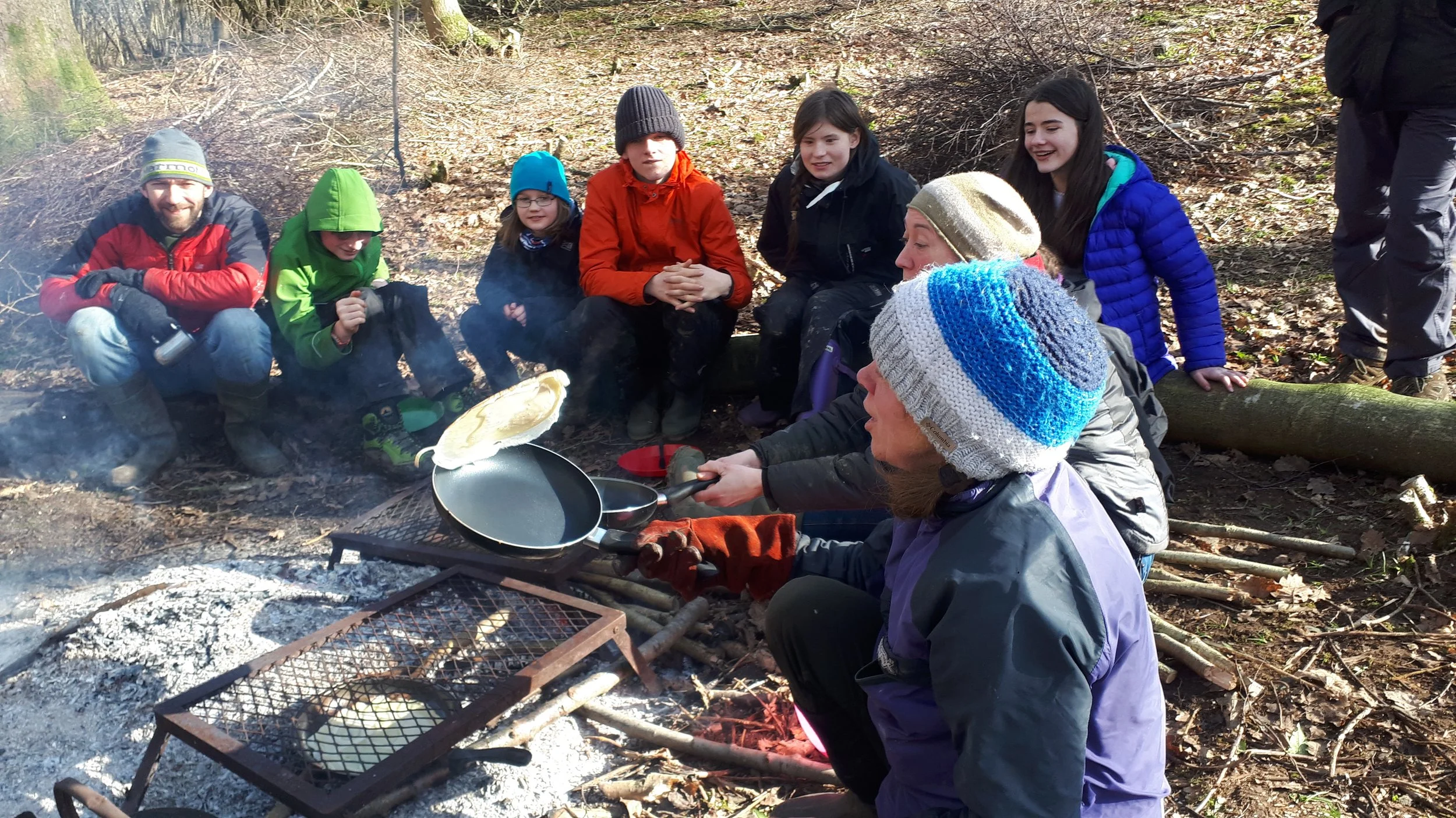 Group of children and adults sitting around a campfire in a wooded area, some wearing winter jackets and hats, with a person cooking over the fire.