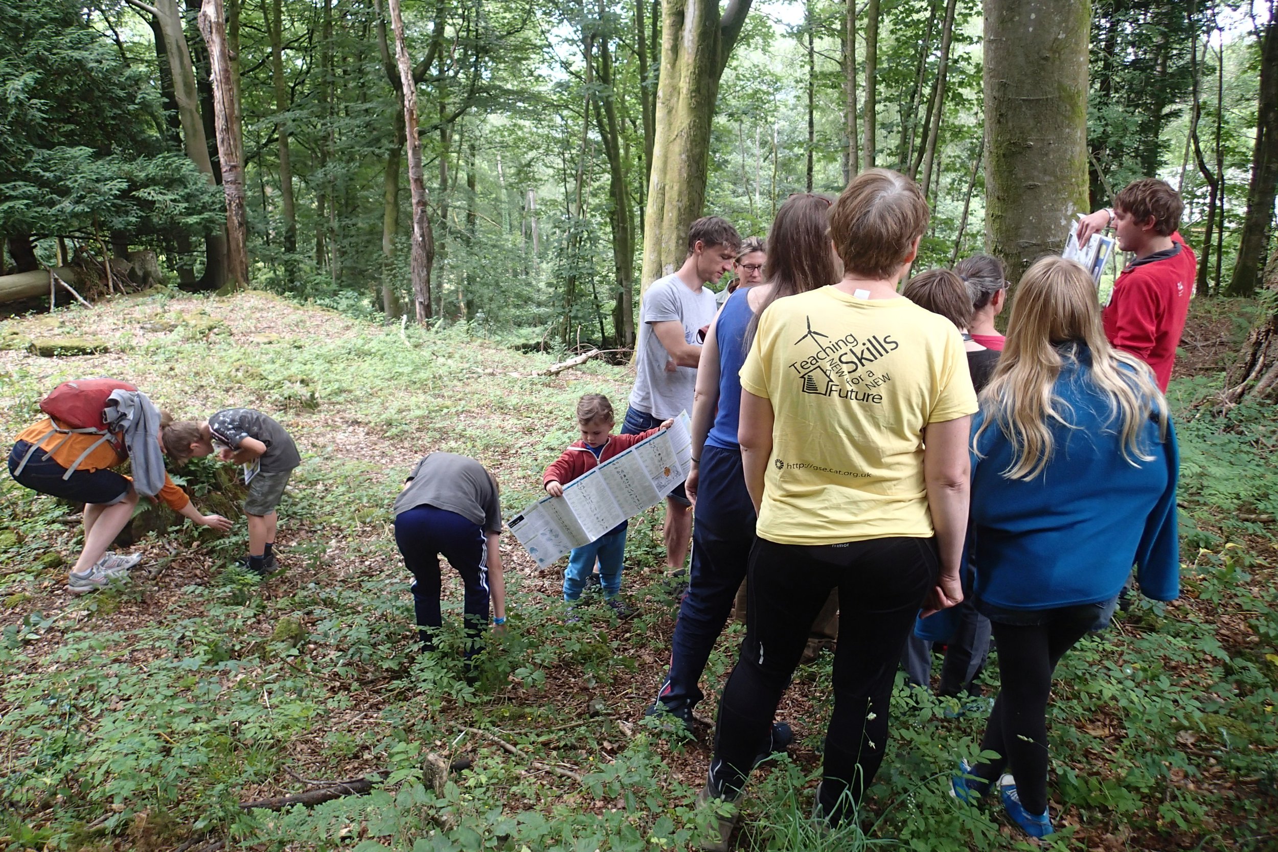 Group of children and teenagers in a forest, some examining plants on the ground, others holding maps, and a guide presenting information in a wooded area.