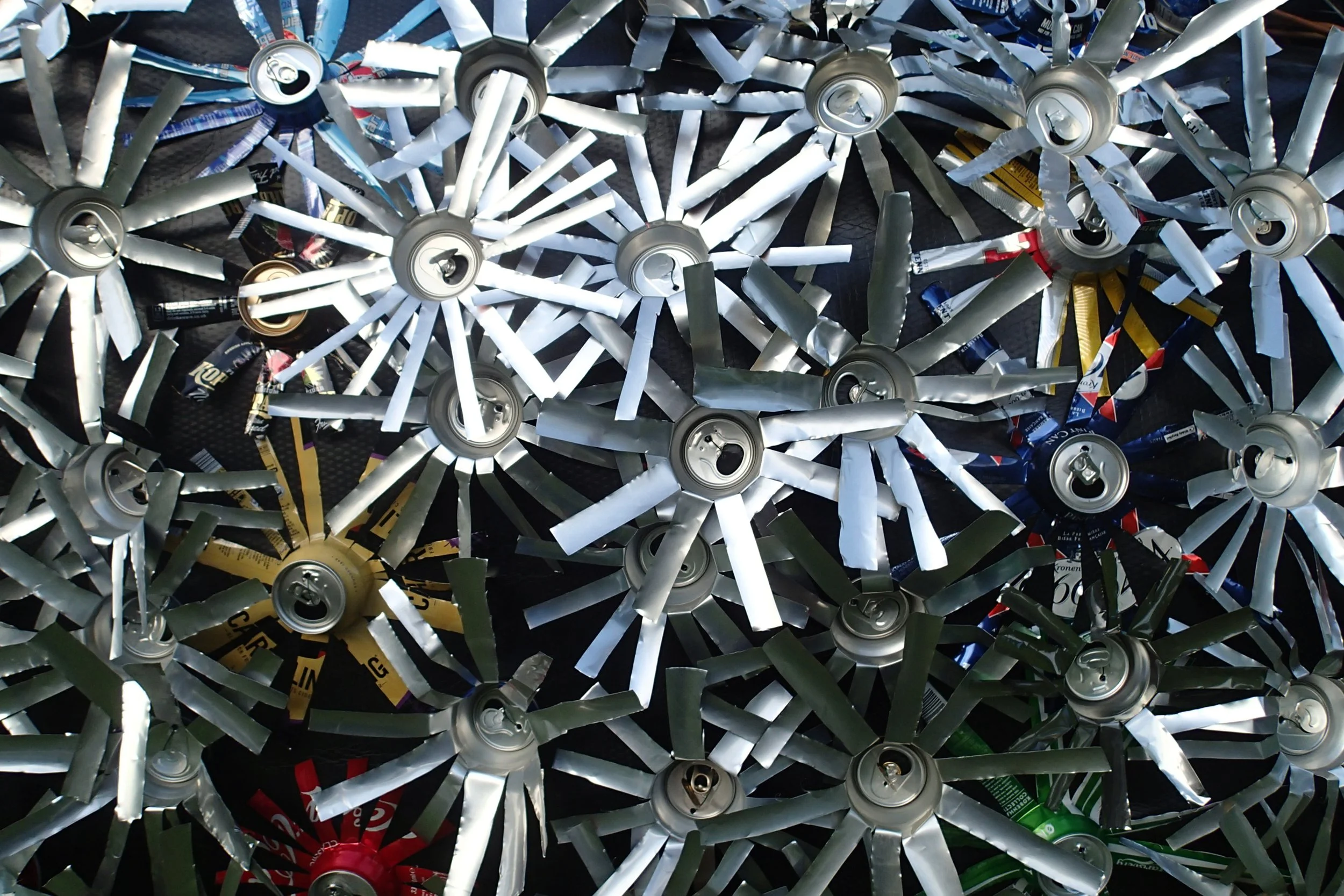 A collection of crushed aluminum cans arranged with their tops and pull-tabs visible, in various colors including silver, red, green, black, blue, and yellow.