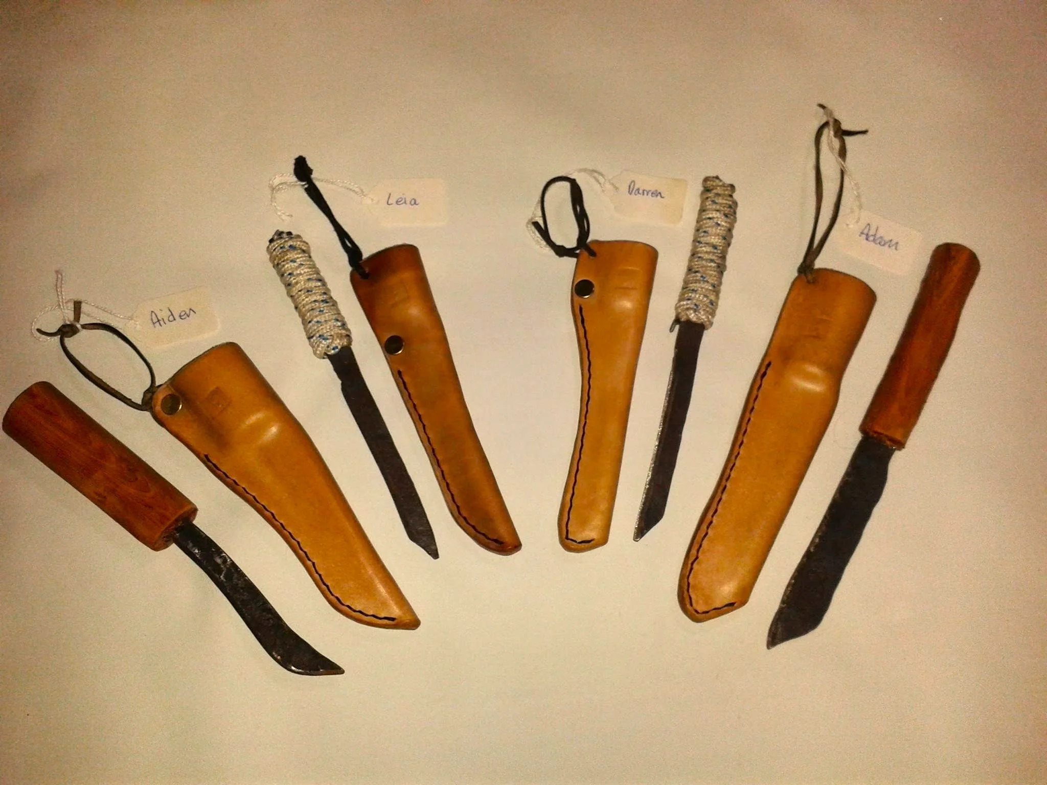 Six knives with leather or wood handles, each with a handwritten name tag. The knives have different blade shapes and sizes, with some handles wrapped in cord or woven material.