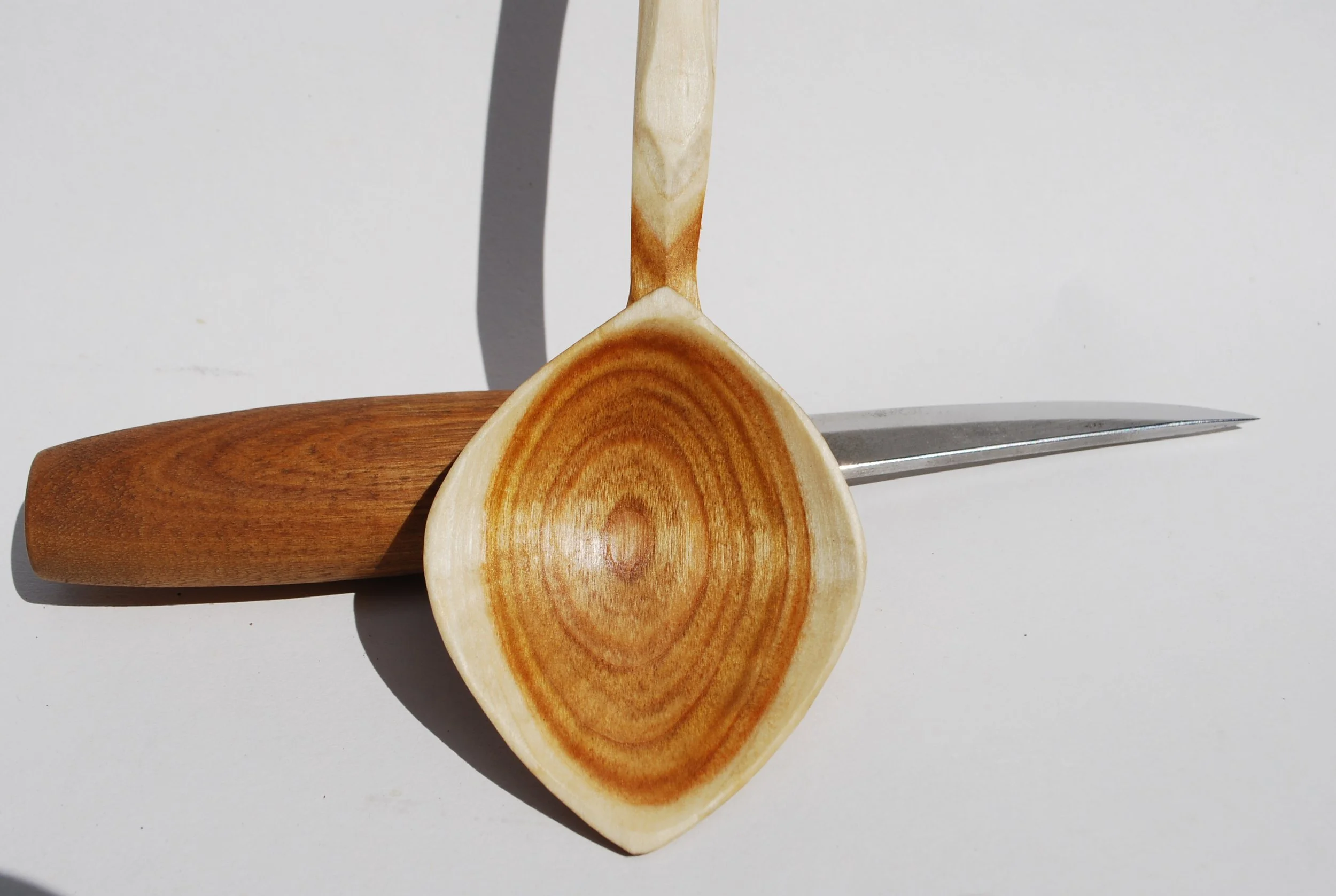 A wooden spoon, a knife, and a wooden spatula arranged on a white surface with shadows.