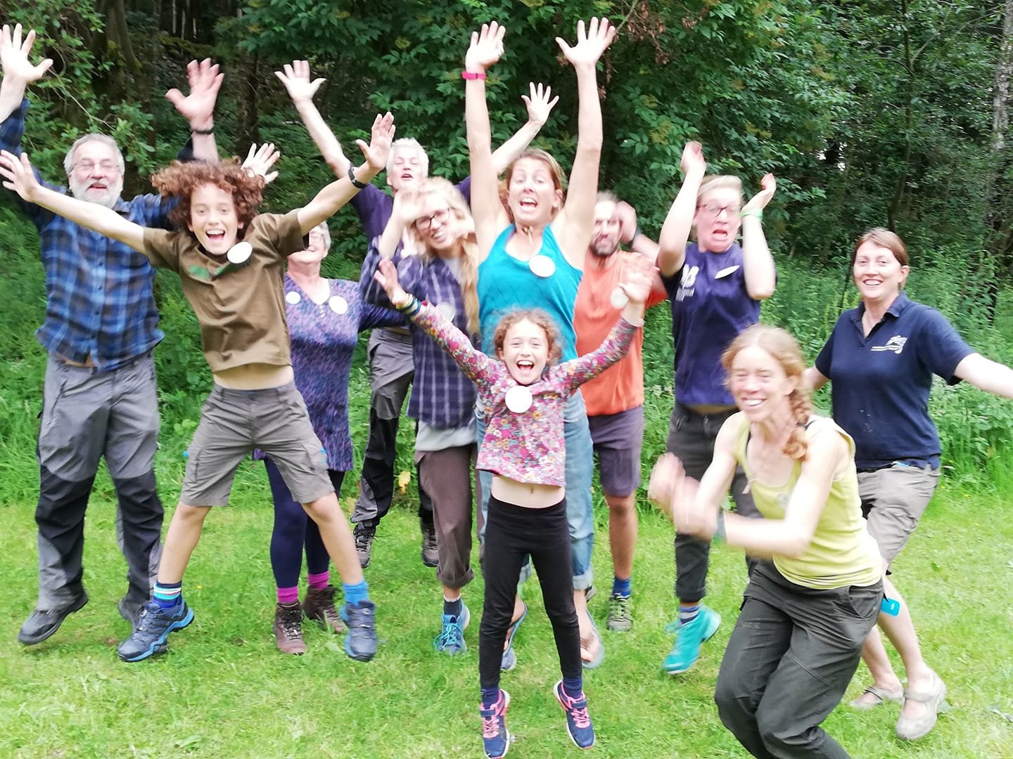 Group of smiling people jumping outdoors on green grass with trees in background.