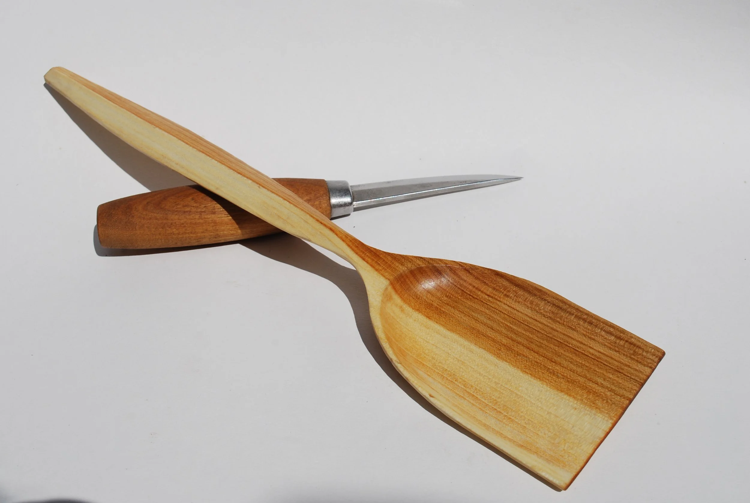 A wooden spoon, a wooden-handled carving tool, and a metal awl arranged on a white surface.