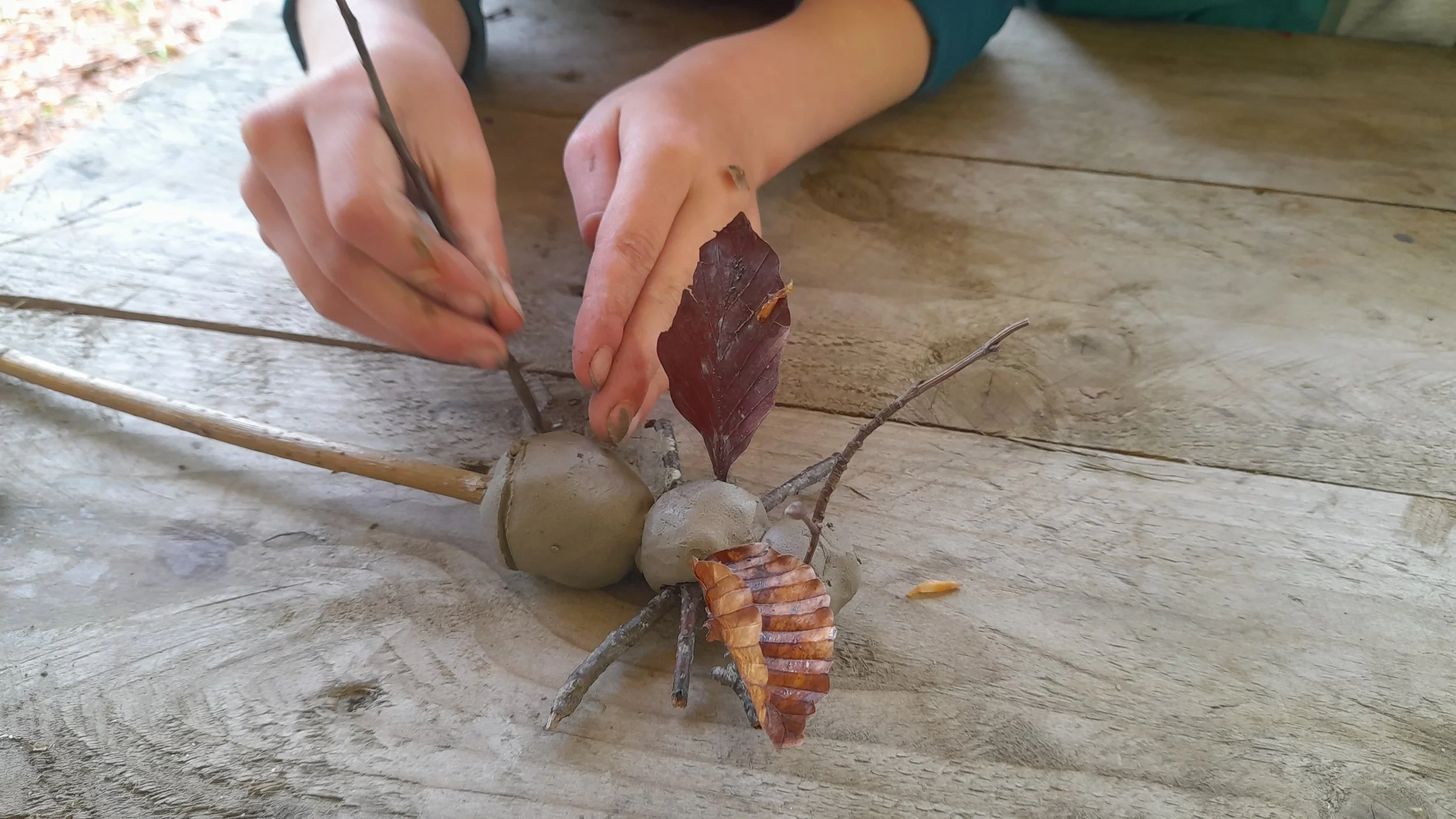 Person using clay to create a sculpture resembling a bee, with sticks, leaves, and other natural materials, on a wooden surface.