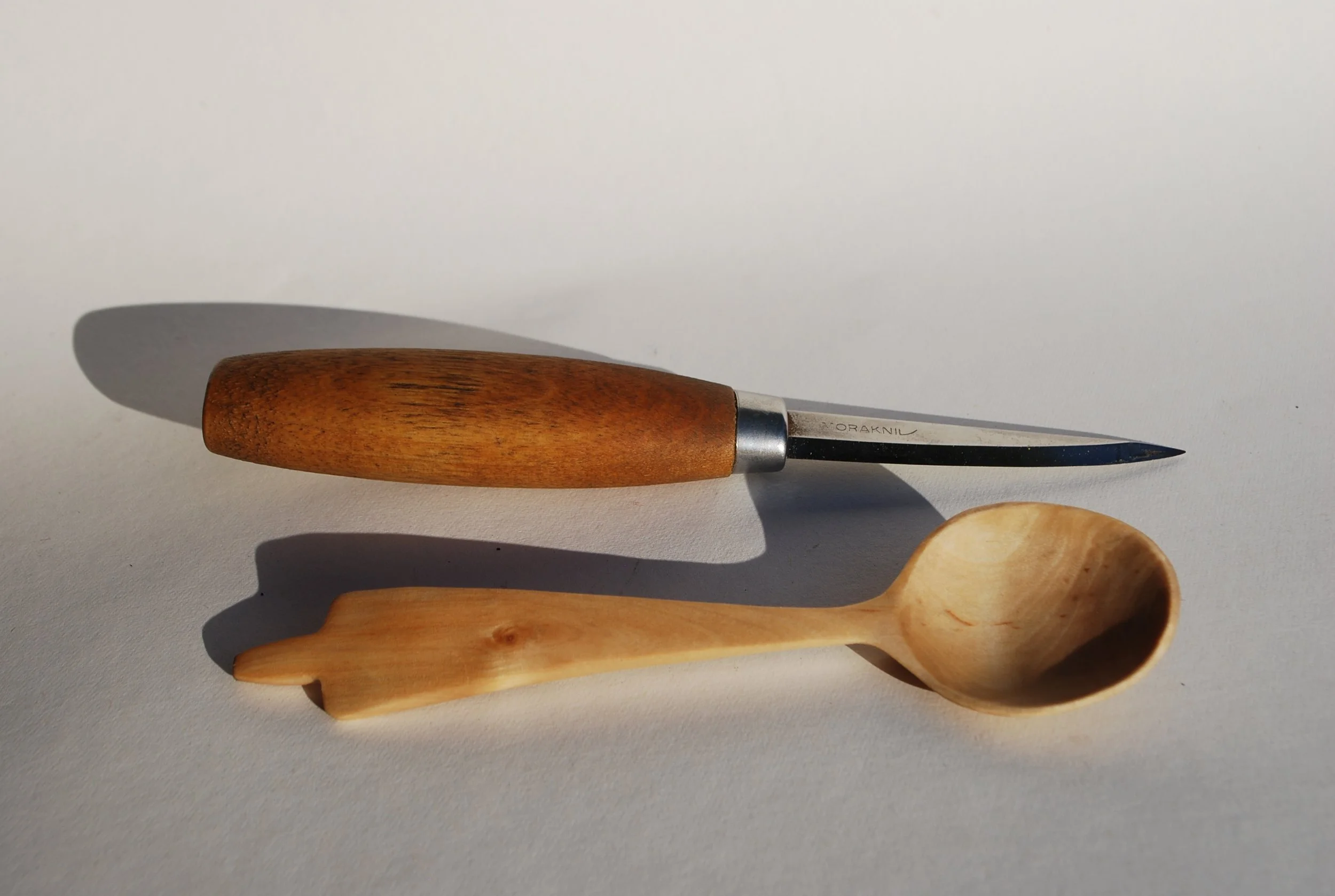 A wood-handled paring knife and a small wooden spoon on a white surface with shadows.