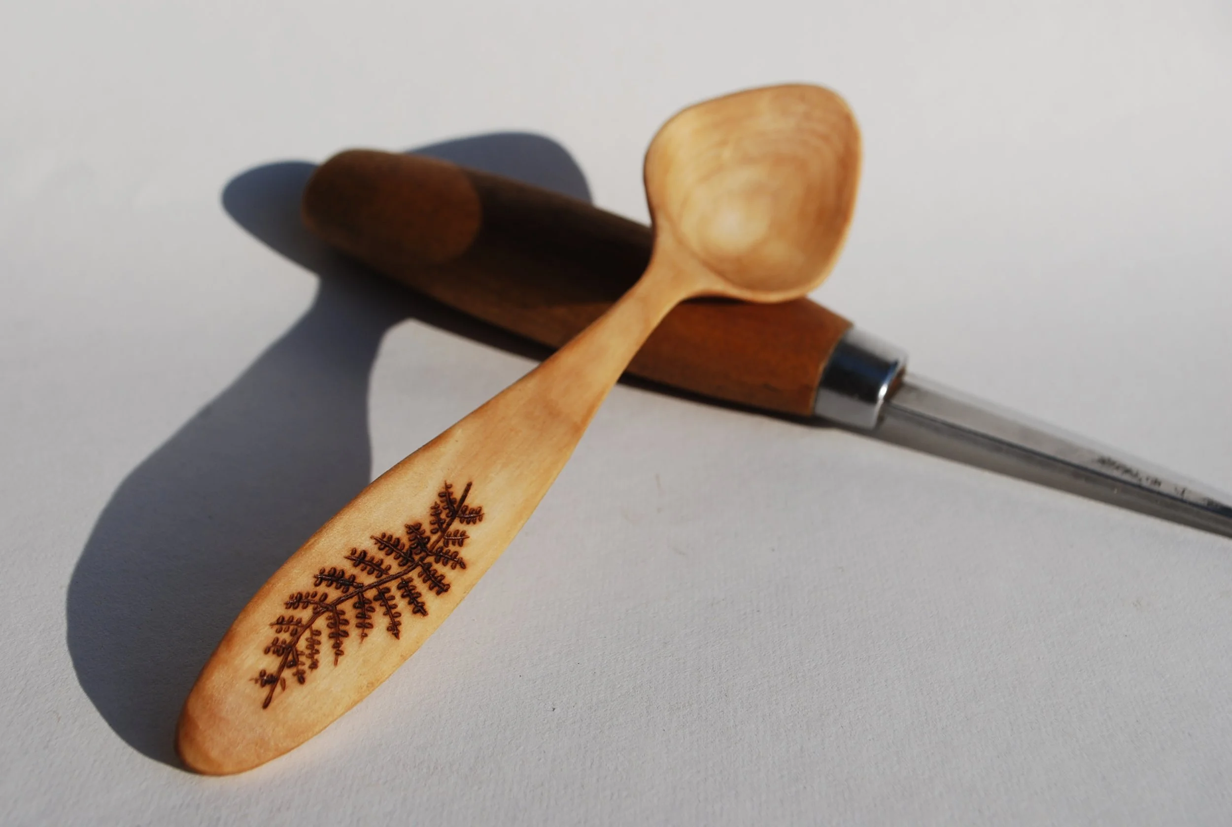 A wooden spoon with a decorative leaf design, a wooden kitchen utensil, and a metal-handled glass straw, all placed on a white surface.