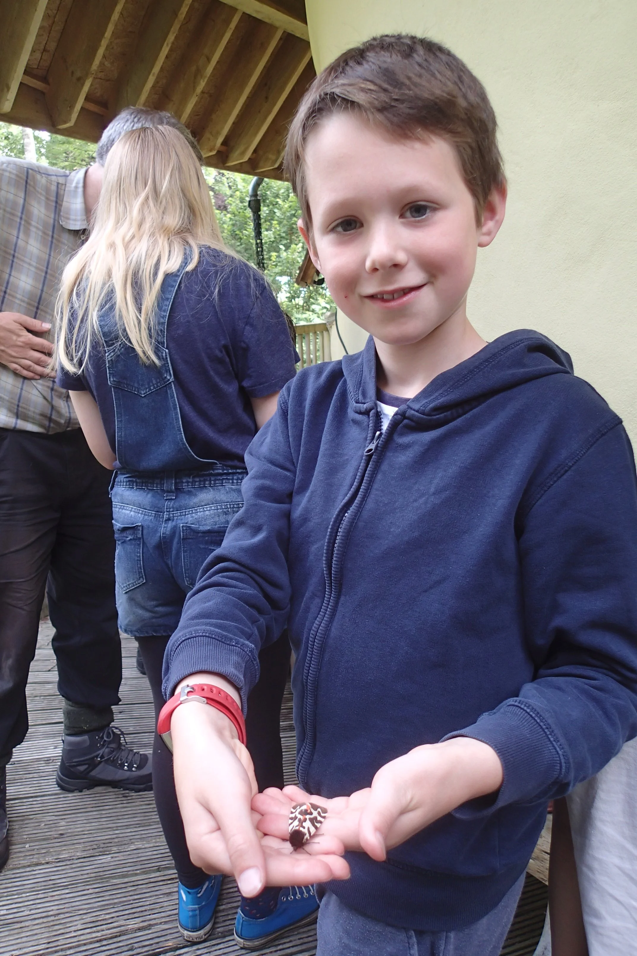 A young boy in a blue hoodie smiling and holding a small decorated object in his hands, with a group of people in the background.