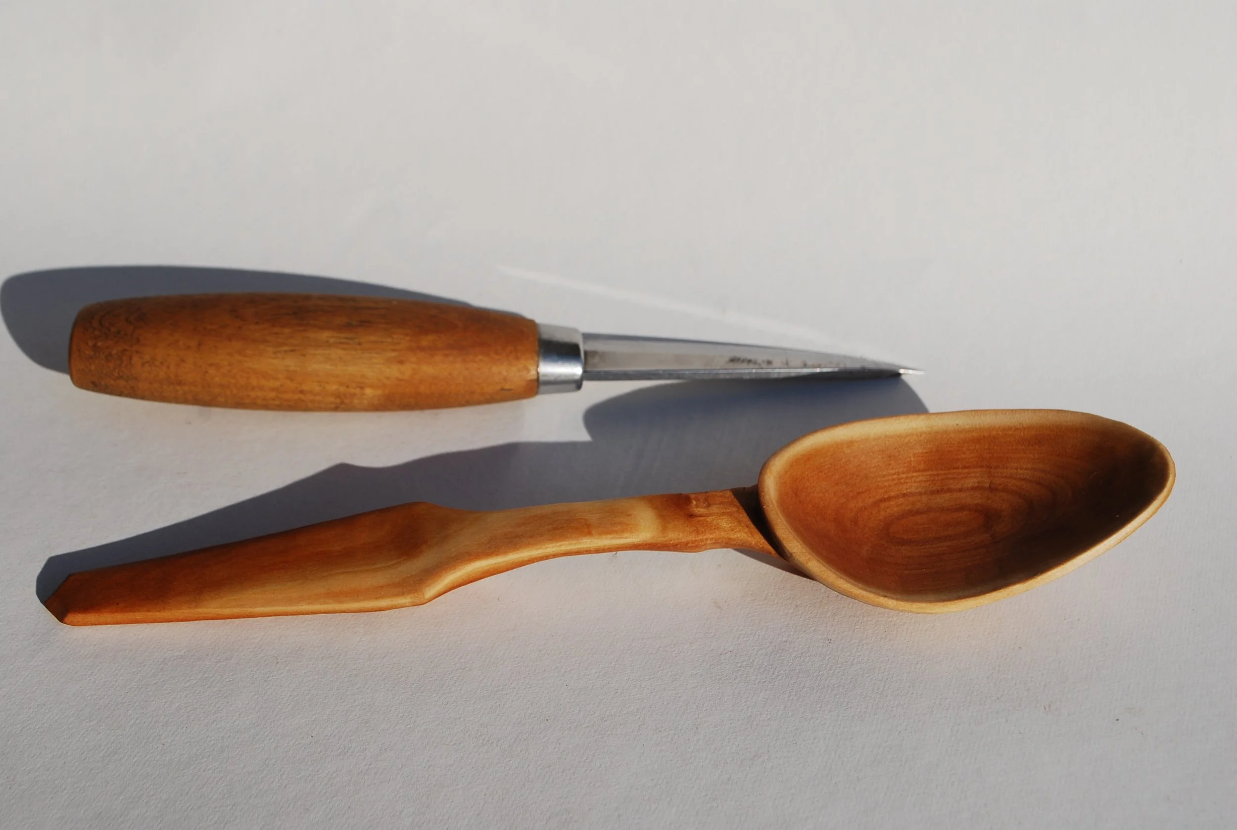 A wooden scoop and a metal and wood kitchen utensil with a rounded end, placed on a white surface with shadows.