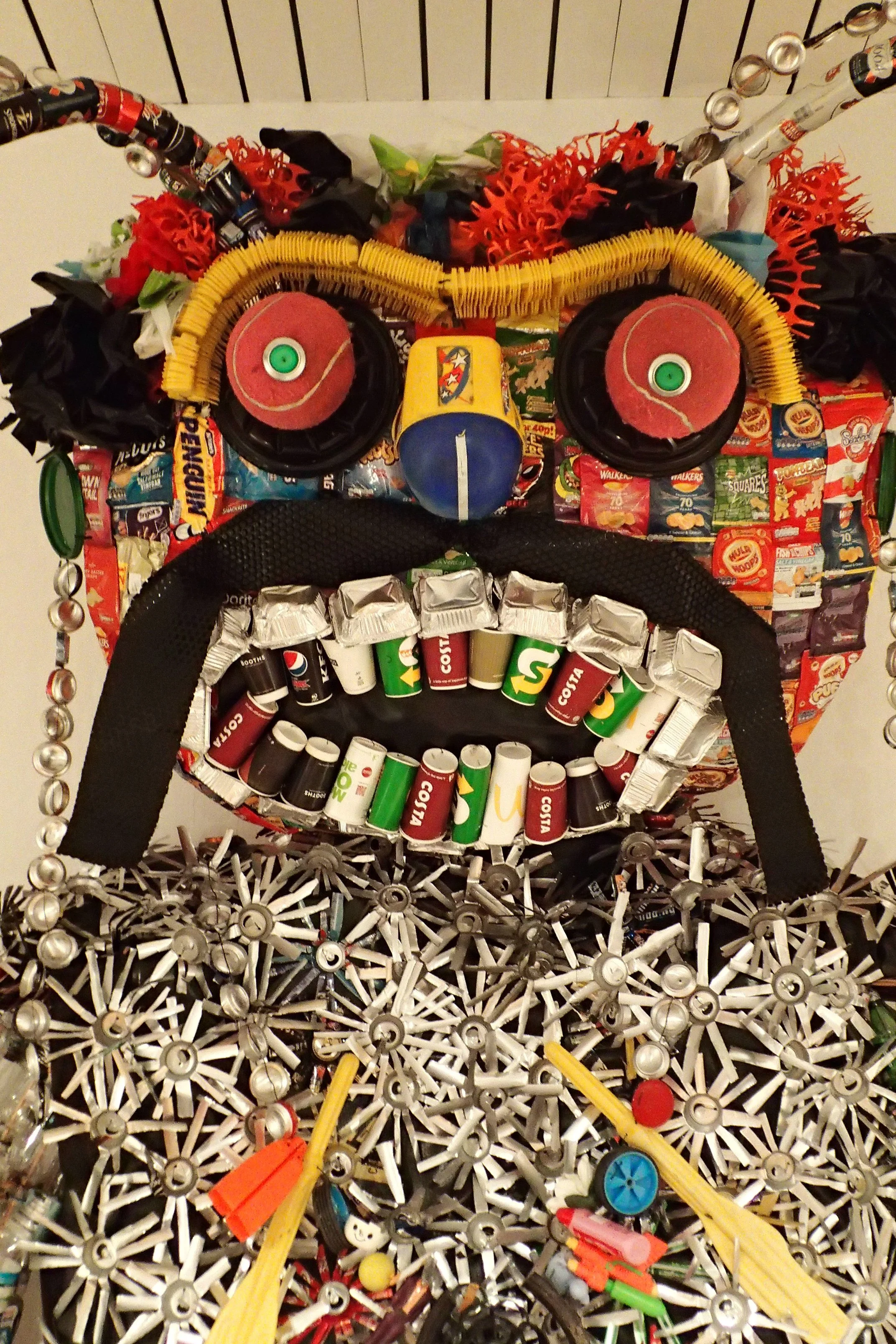 Artistic face created using various recycled materials, including bottle caps, aluminum cans, toy parts, and colorful objects, arranged to form eyes, a nose, and a mouth.