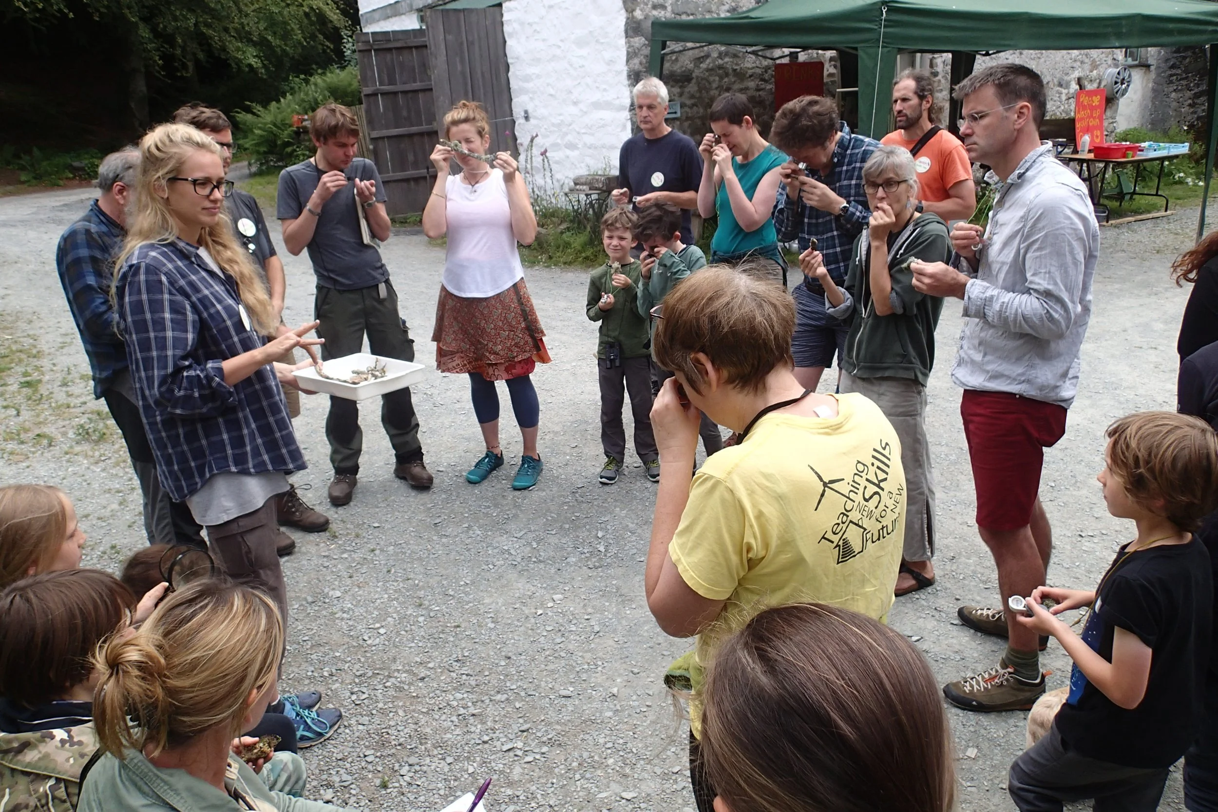 Group of people gathered outdoors, some holding microscopes, with a woman showing a tray of specimens, under a green canopy, on a gravel surface with a white building in the background.