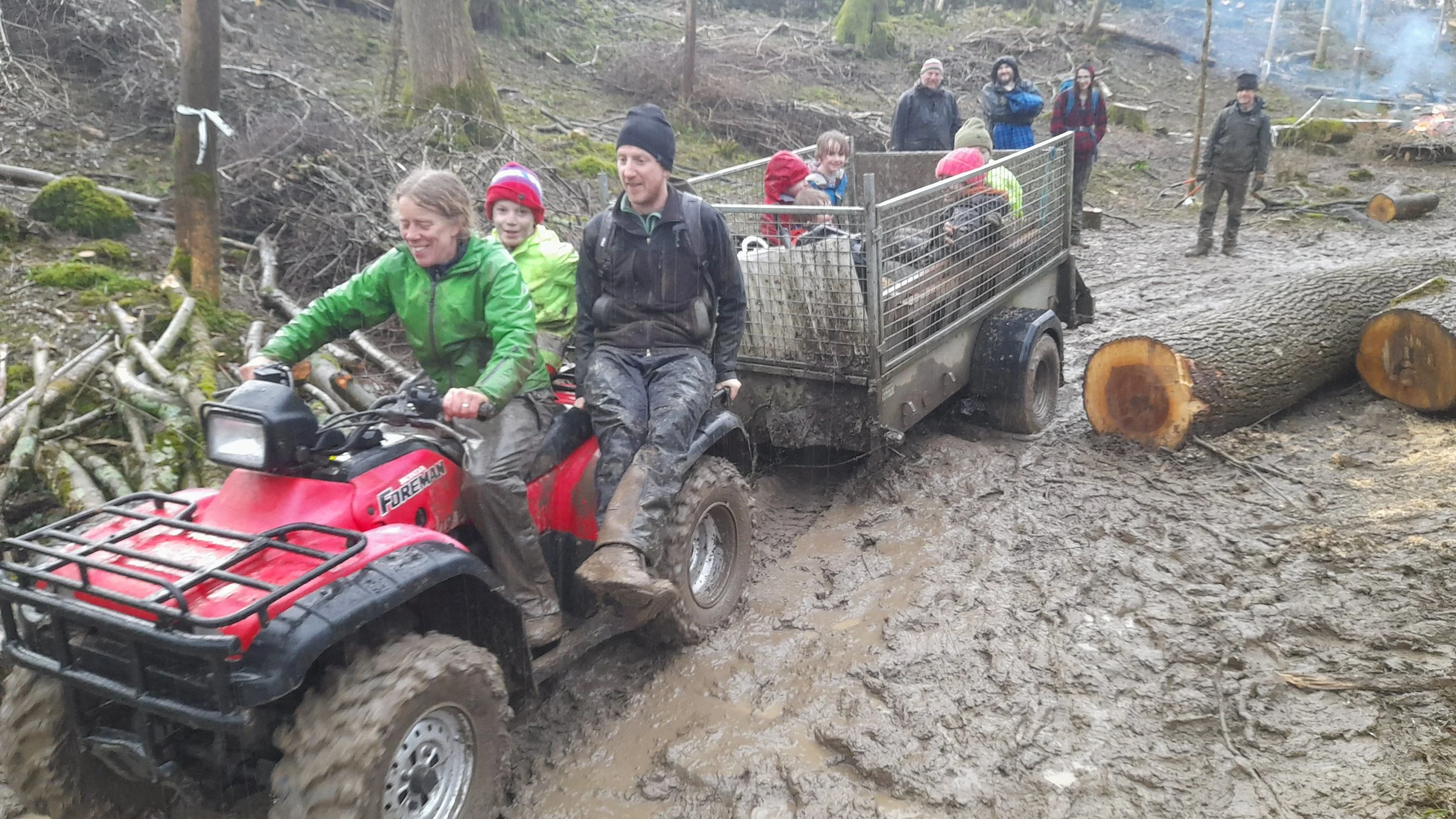 People riding a red ATV with a trailer attached through a muddy forest trail, with others walking and watching in the background.