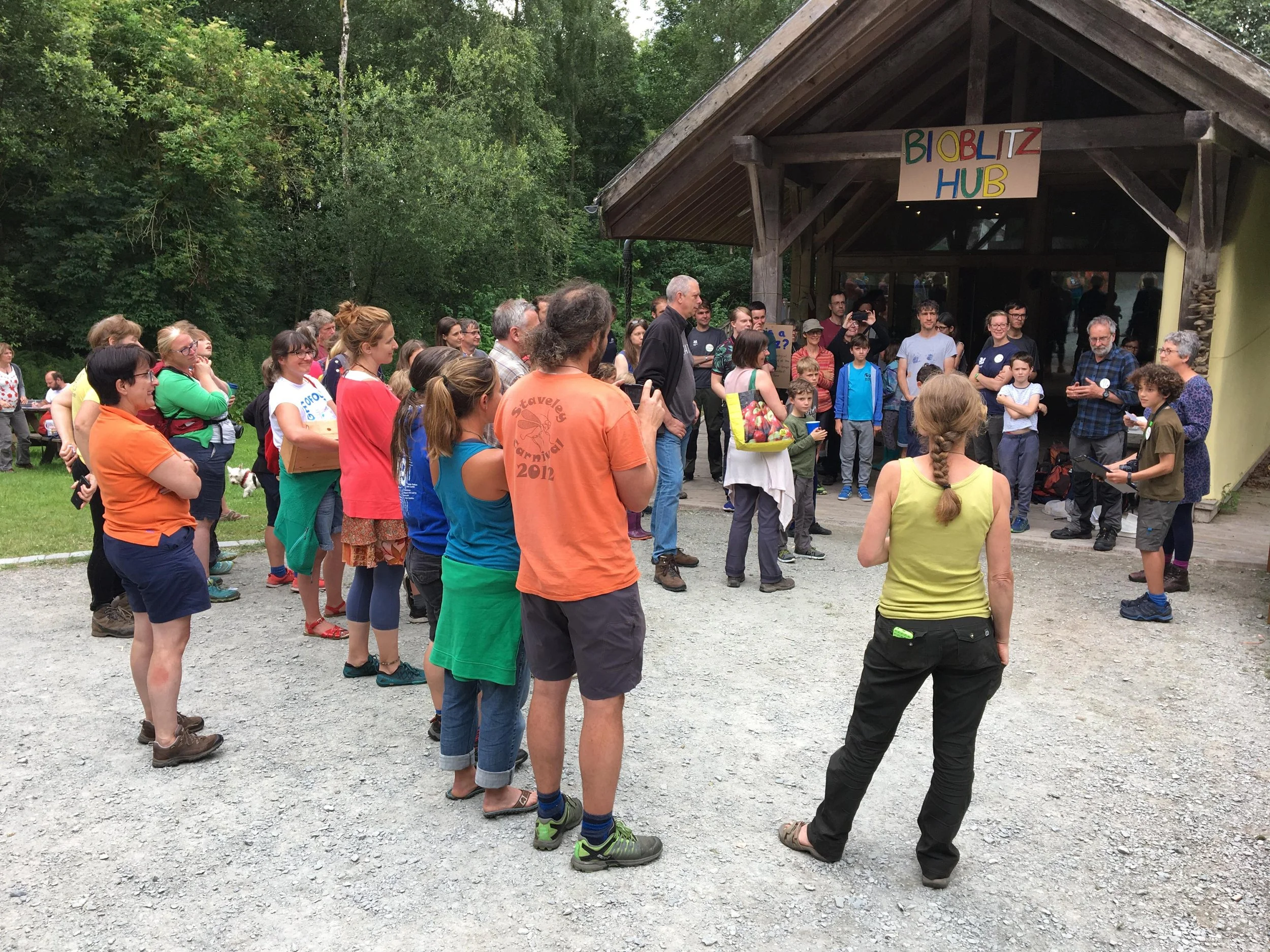 A group of people gathered outside a building with a sign that reads 'BioBlitz Hub,' listening to a speaker in front of the entrance while standing on a gravel surface, with trees in the background.