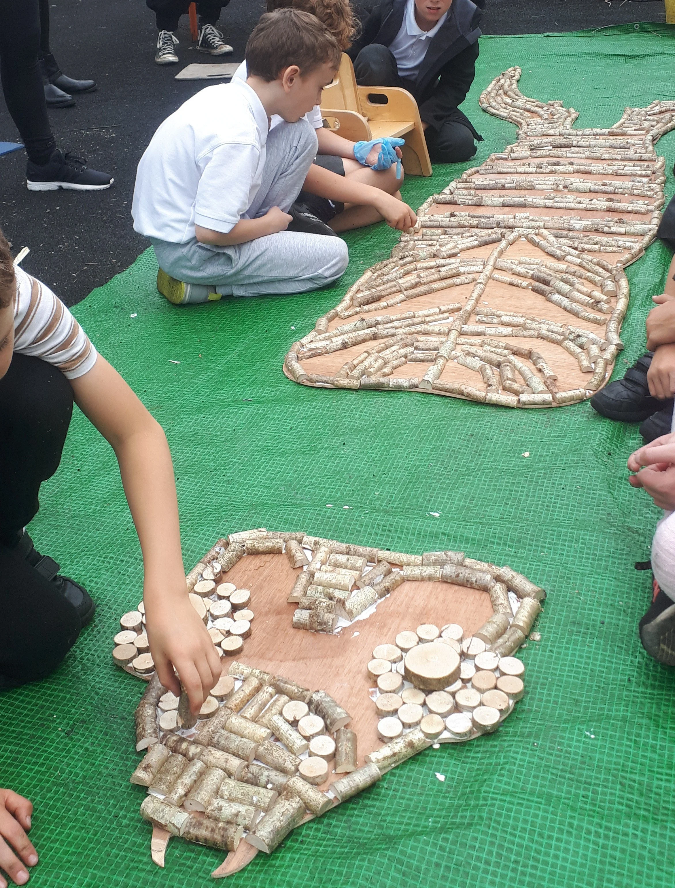 Children laying out and assembling a large mosaic or art project made of small logs and wood slices on a green tarp outdoors.