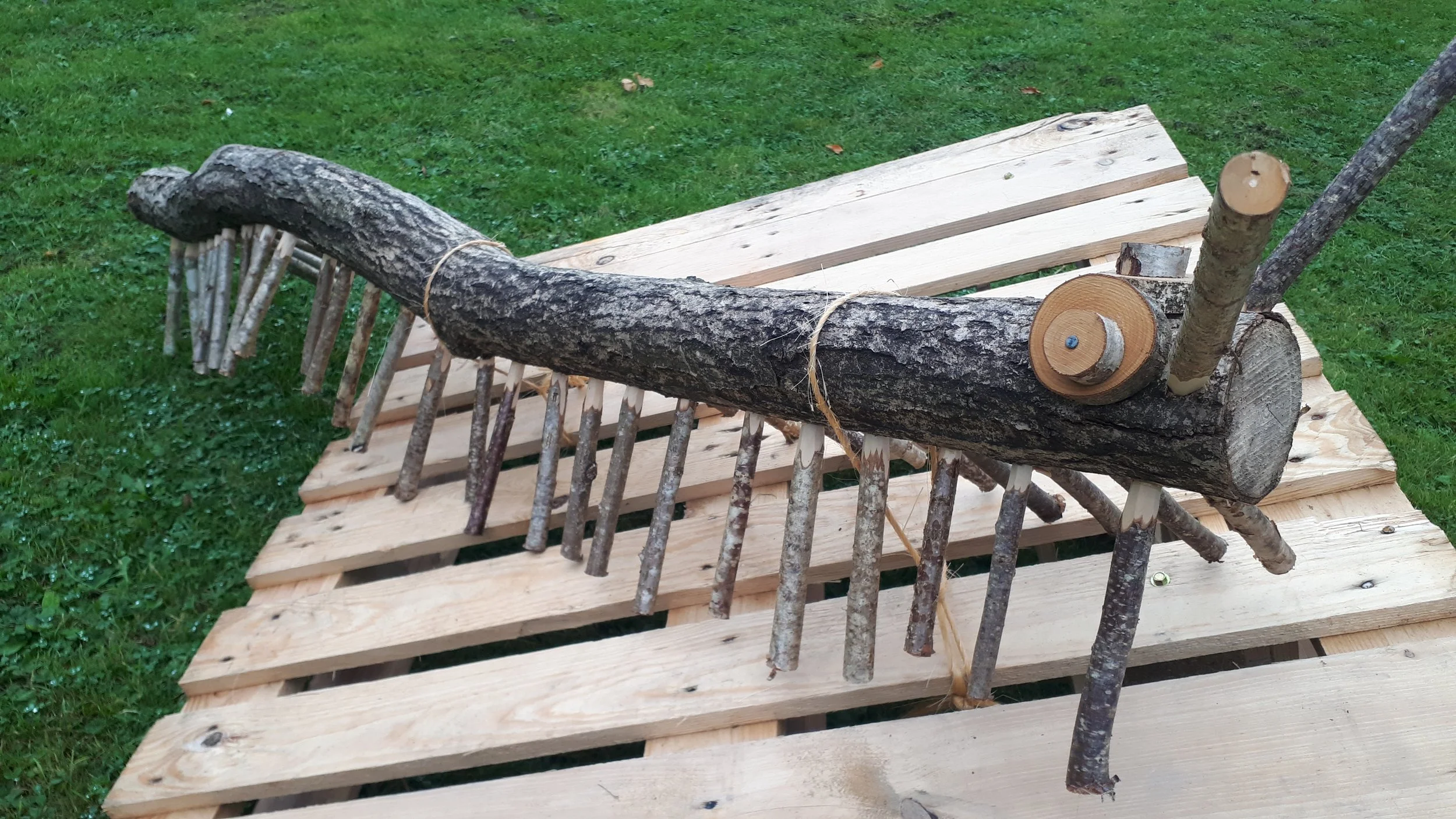A wooden rake made from natural sticks and branches, placed on a stack of light-colored wooden pallets on grass.