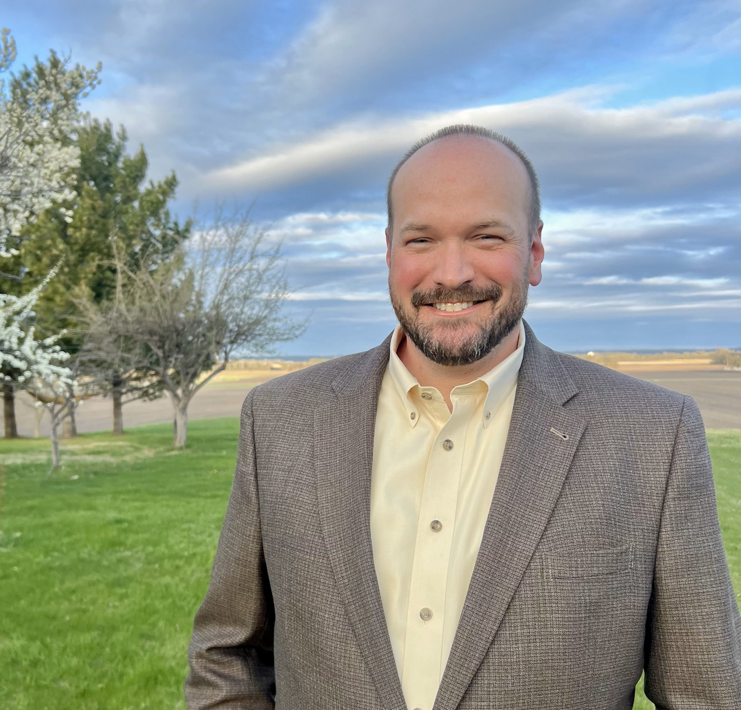 Smiling man in a suit standing on green grass with trees and a cloudy sky in the background.