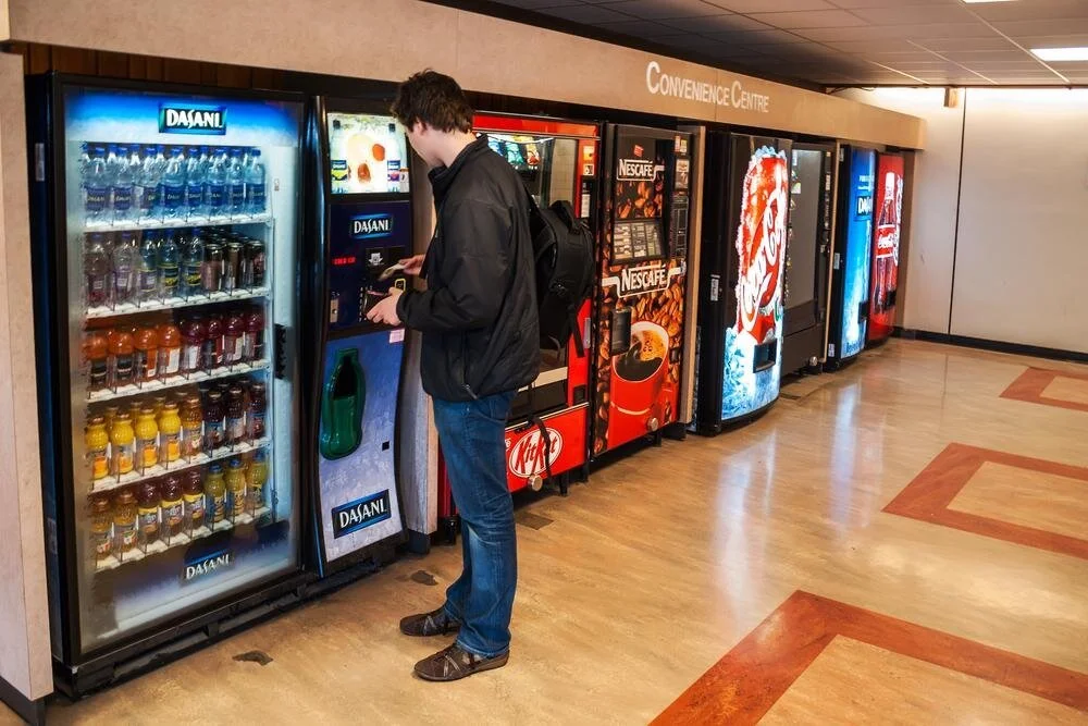 A man with a black backpack buying a bottled beverage from a vending machine in a convenience center.