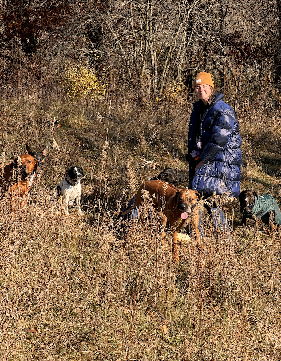 A woman with short hair, wearing a yellow beanie and a blue puffy coat, crouching in a grassy field with five dogs around her. The scene is outdoors with leafless trees in the background, indicating fall or early winter.