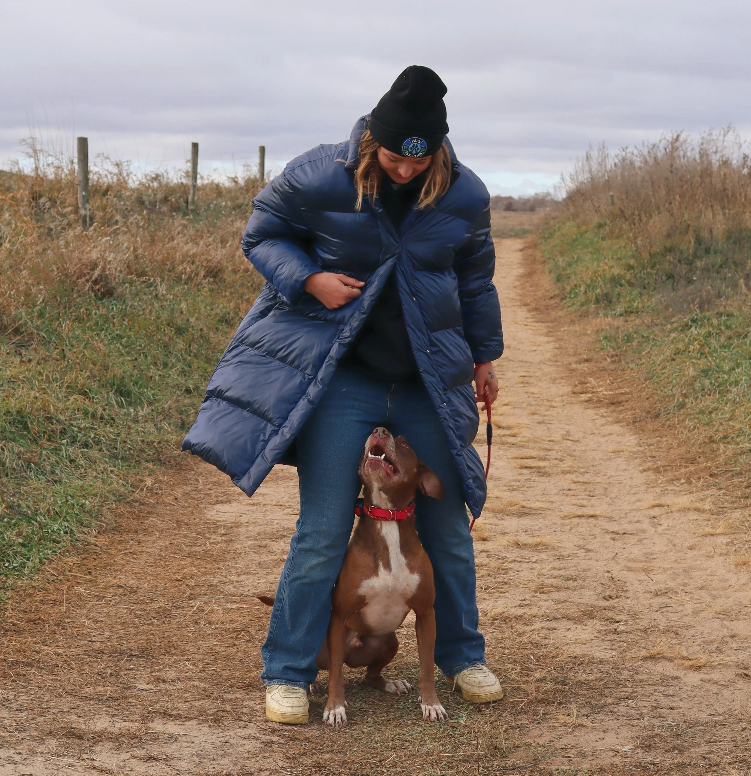 A woman in a black beanie and navy blue winter coat standing on a dirt path, smiling down at a brown dog sitting between her legs, looking up and barking.