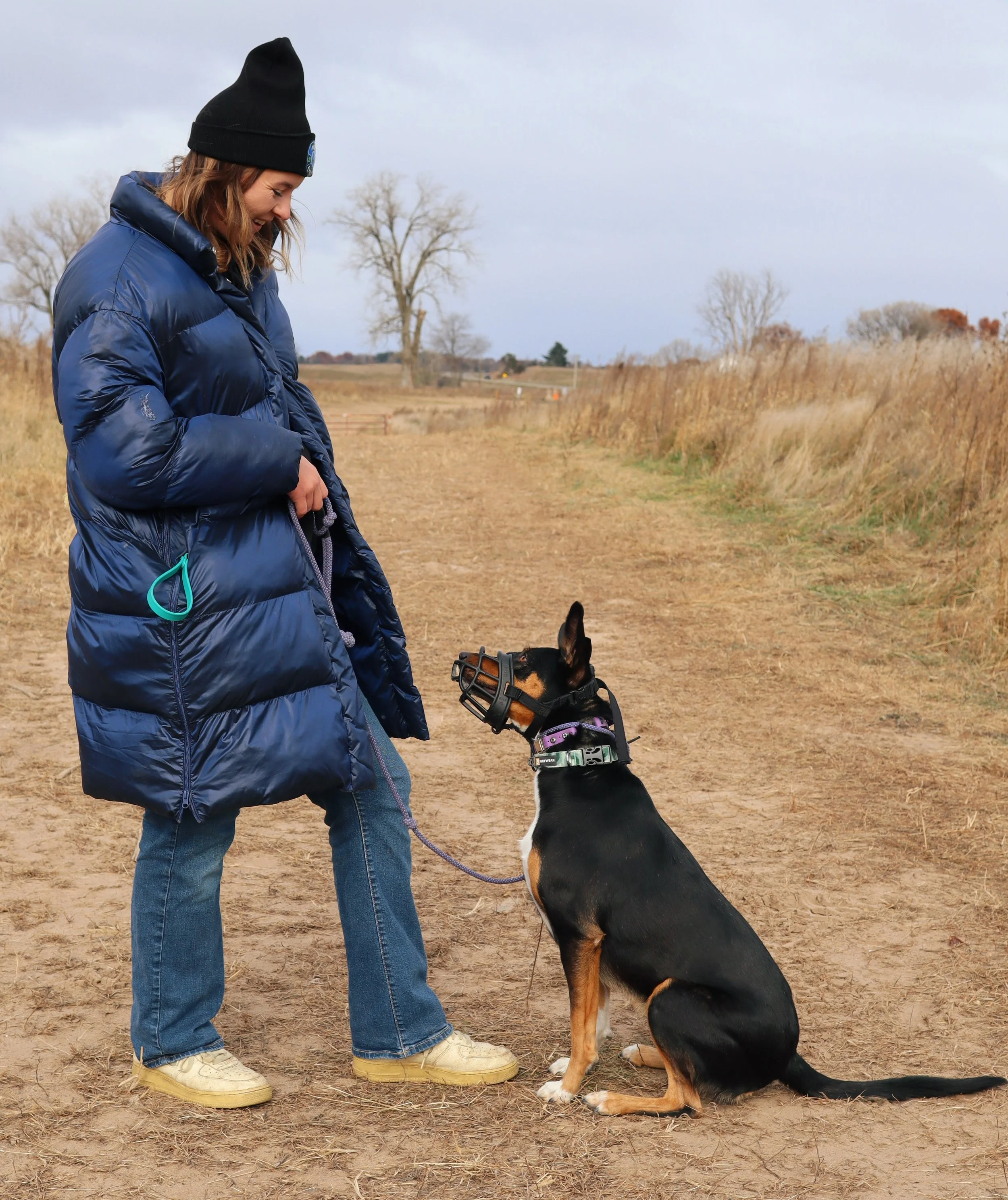 A woman in a puffy blue jacket and black beanie standing on a dirt path with her dog, which is wearing a muzzle and harness, outdoors during daytime.
