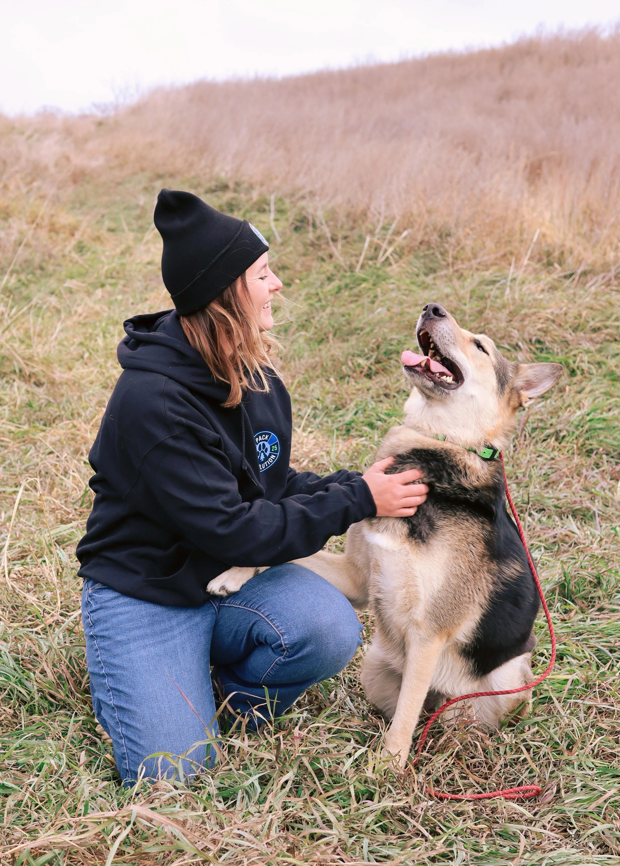 A woman kneeling outdoors on grass, smiling at a happy dog with a tan and black coat, in a grassy field with a hill behind them.