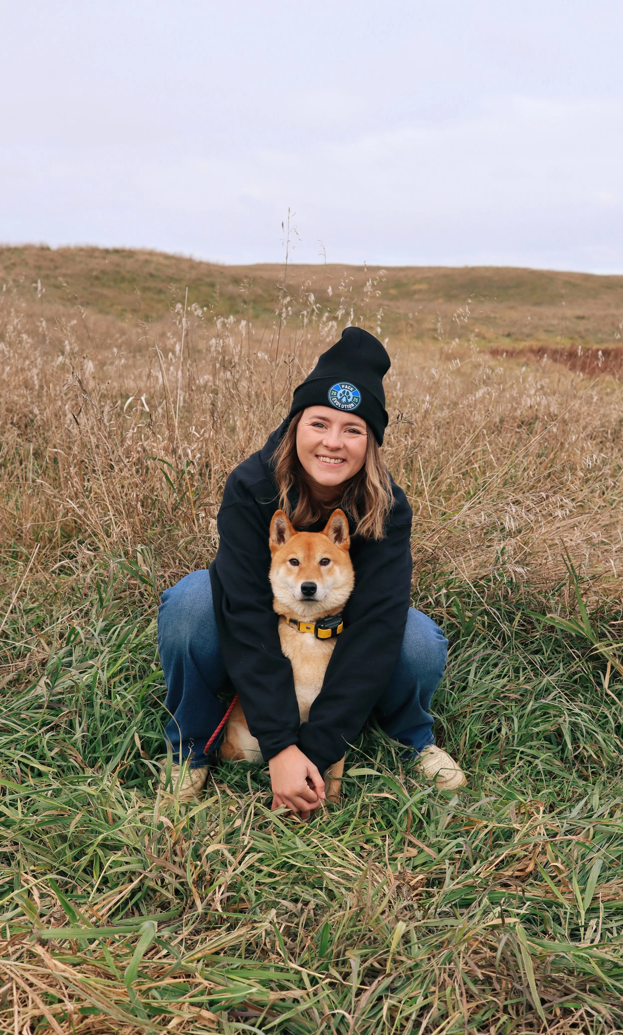 A woman squatting in a field of tall grass with a Shiba Inu dog, both looking at the camera, in an open landscape with rolling hills and a cloudy sky.