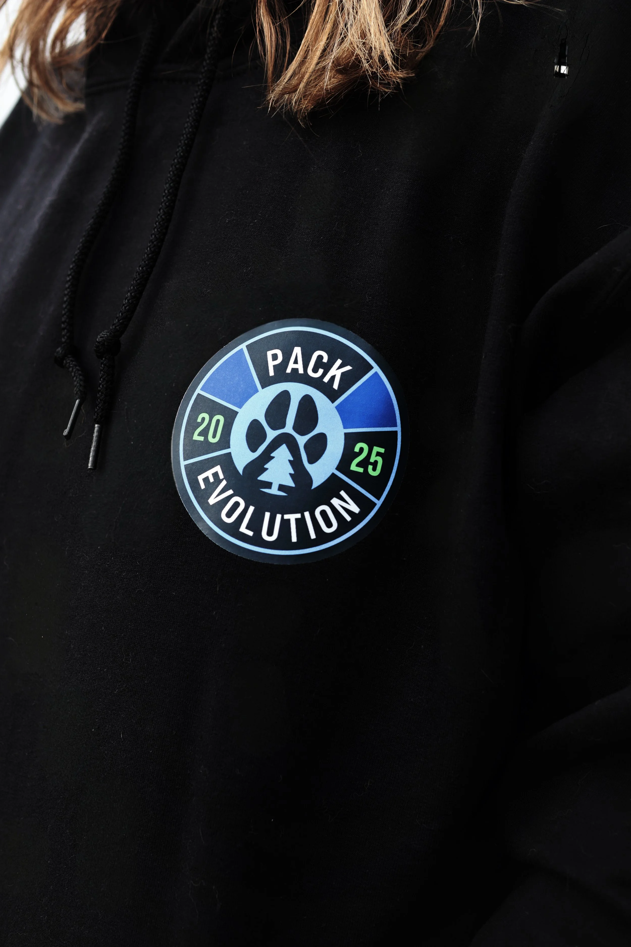 Black hoodie with a circular patch that reads 'Pack Evolution 2025' and features a paw print, tree, and blue and green sections.
