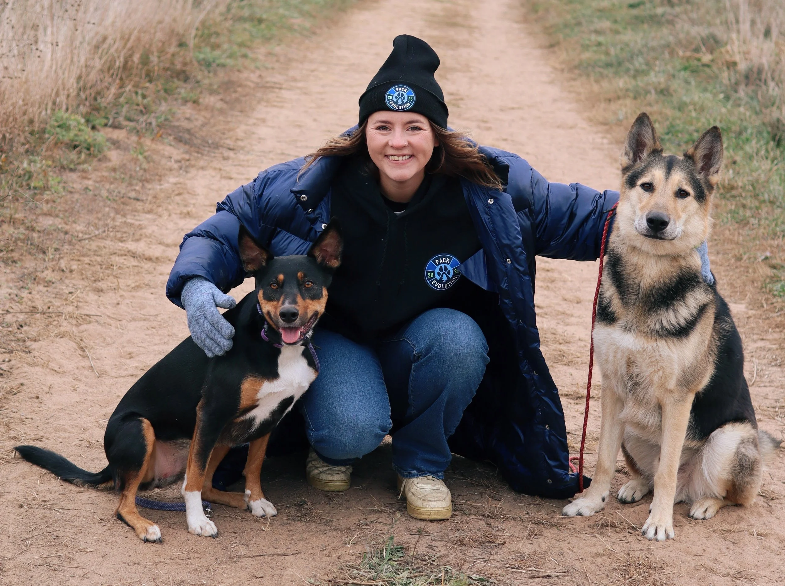 A woman crouching on a dirt path outdoors, smiling, with her arms around two dogs. She is wearing a black beanie, a navy blue puffer jacket, a black hoodie with a 'Pack Evolution' logo, jeans, and gloves. The dog on her left is a small black and tan mixed breed, sitting with its tongue out. The dog on her right is a larger gray and black Siberian Husky mix, sitting and looking at the camera.