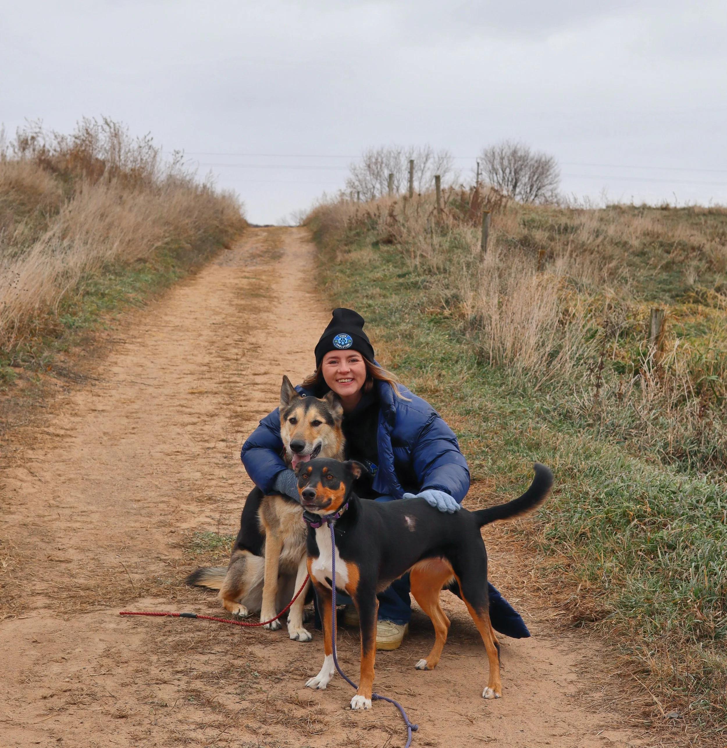 A woman smiling and hugging two dogs on a dirt trail in a rural area with dry grass and overcast sky.