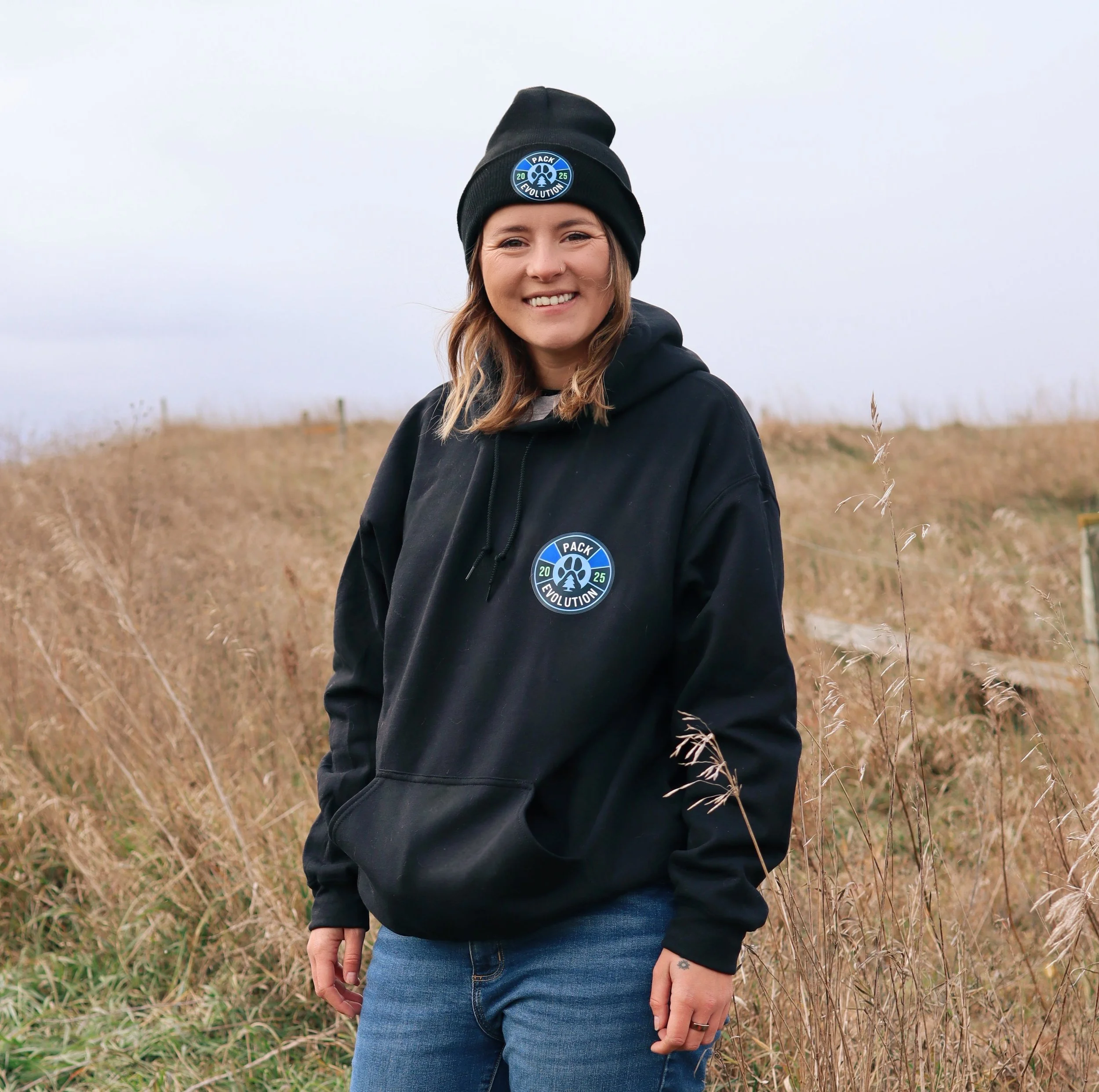 A young woman standing outdoors in a field of tall, dry grass with a cloudy sky. She is wearing a black beanie and a black hoodie with a circular logo that says 'PACK EVOLUTION 2025' and smiling at the camera.