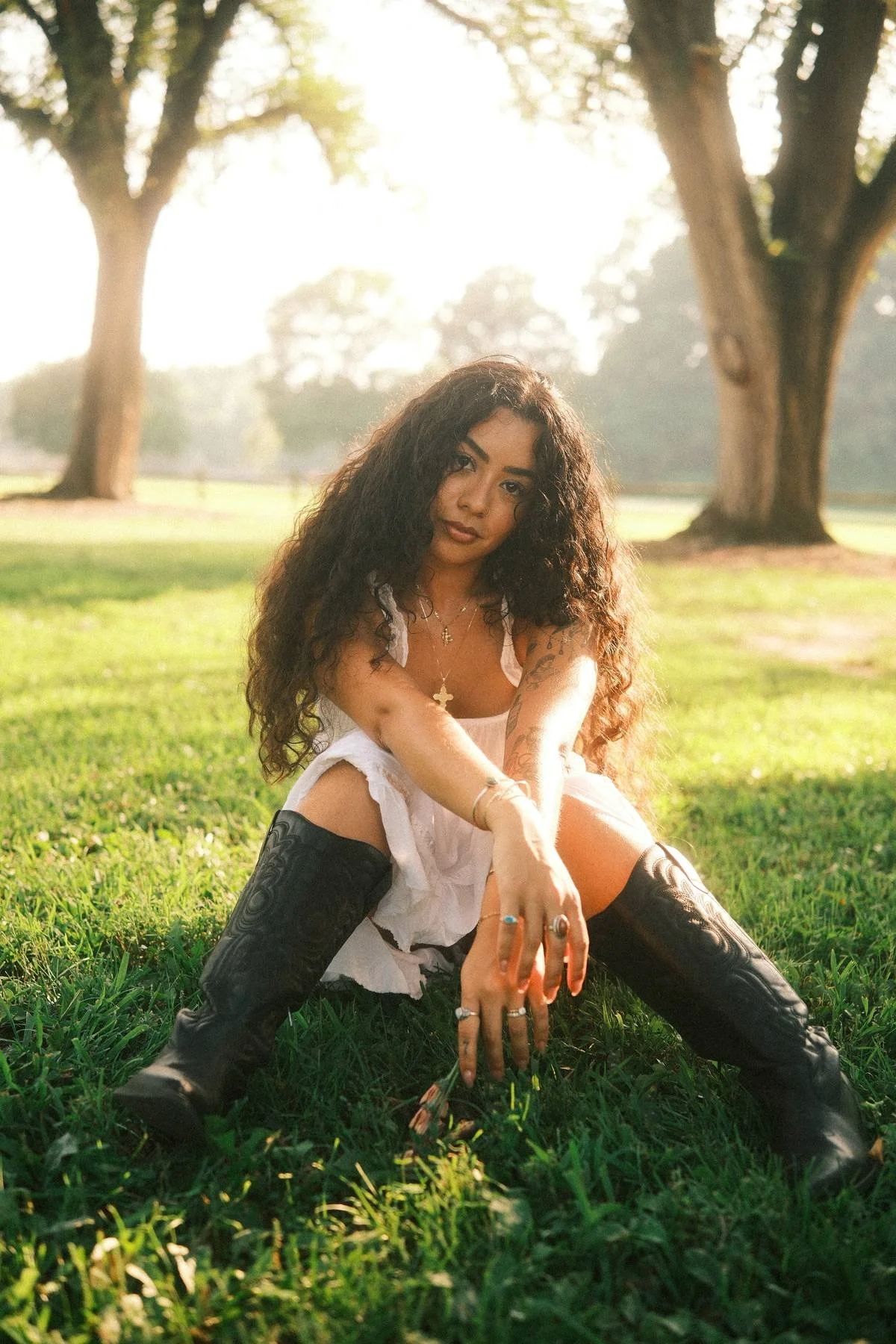 A young woman with long, curly hair sitting on grass in a park with trees in the background. She is wearing a white dress, black knee-high boots, and various jewelry. Sunlight filters through the trees, creating a warm, glowing atmosphere.
