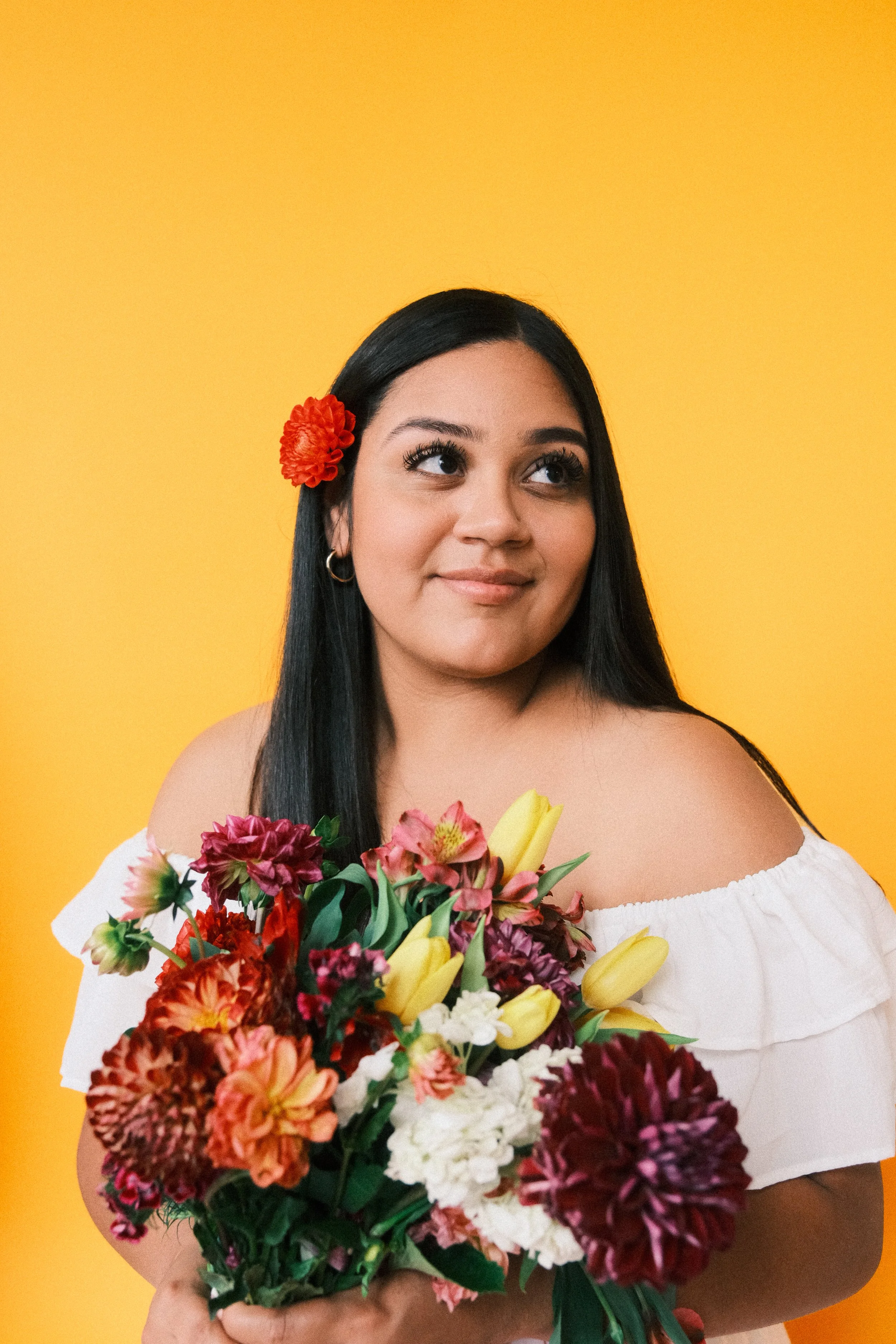 A woman with long black hair, wearing a white off-the-shoulder top and a red flower in her hair, holding a colorful bouquet of flowers against a yellow background.
