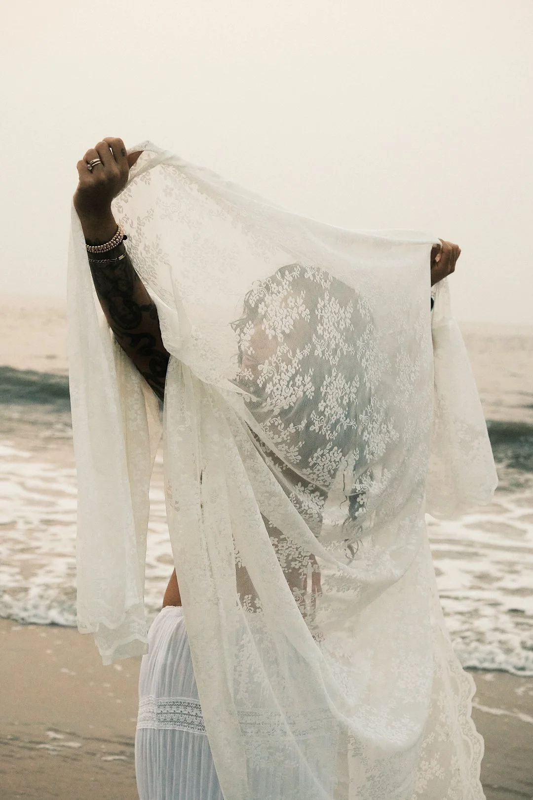 A woman holding a sheer lace fabric in front of her face on the beach at sunset, with ocean waves in the background.
