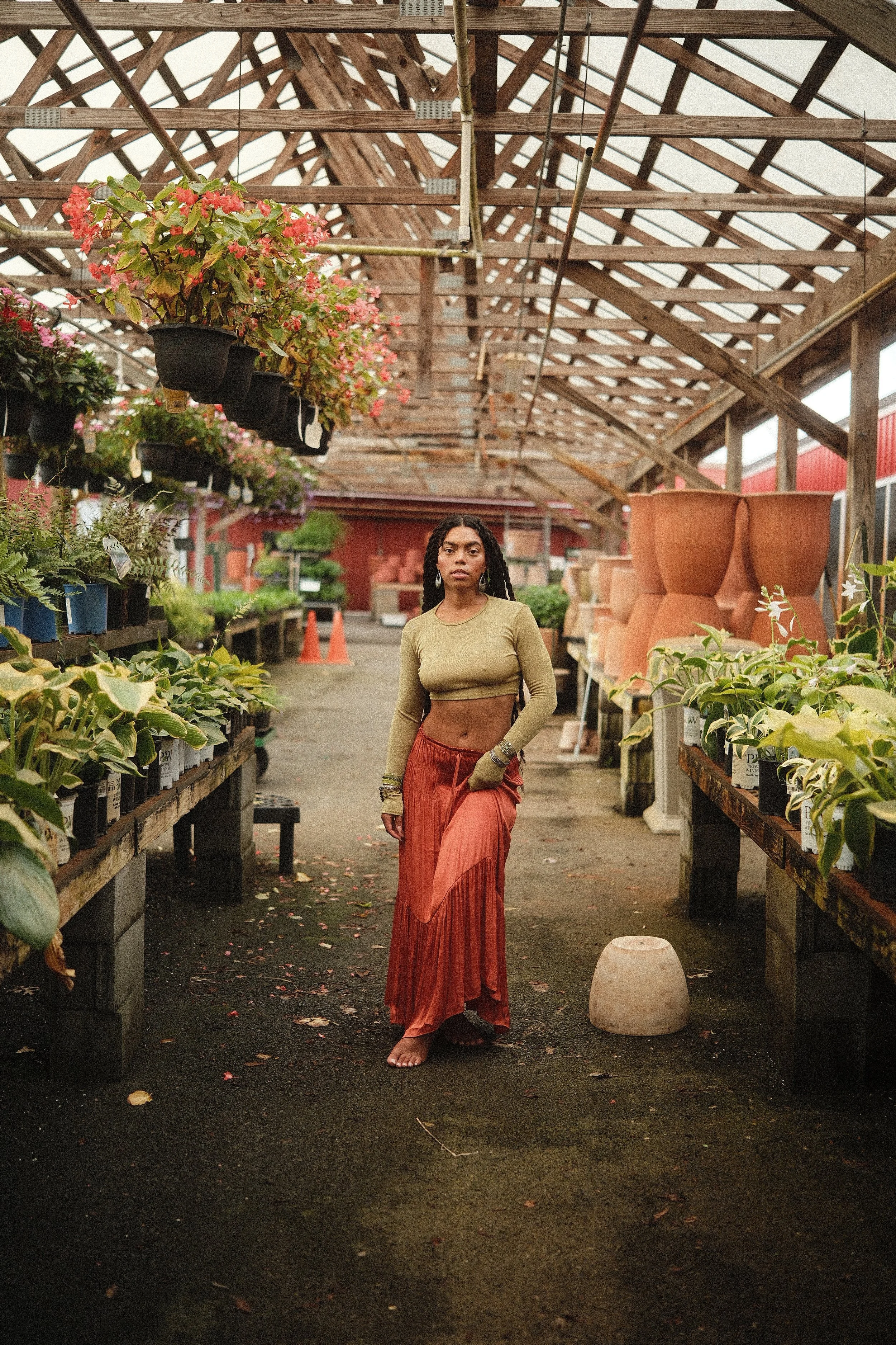 A woman with long dark hair stands barefoot in a greenhouse aisle, wearing a yellow long-sleeve crop top and a flowing orange skirt, surrounded by potted plants and flowers on both sides.
