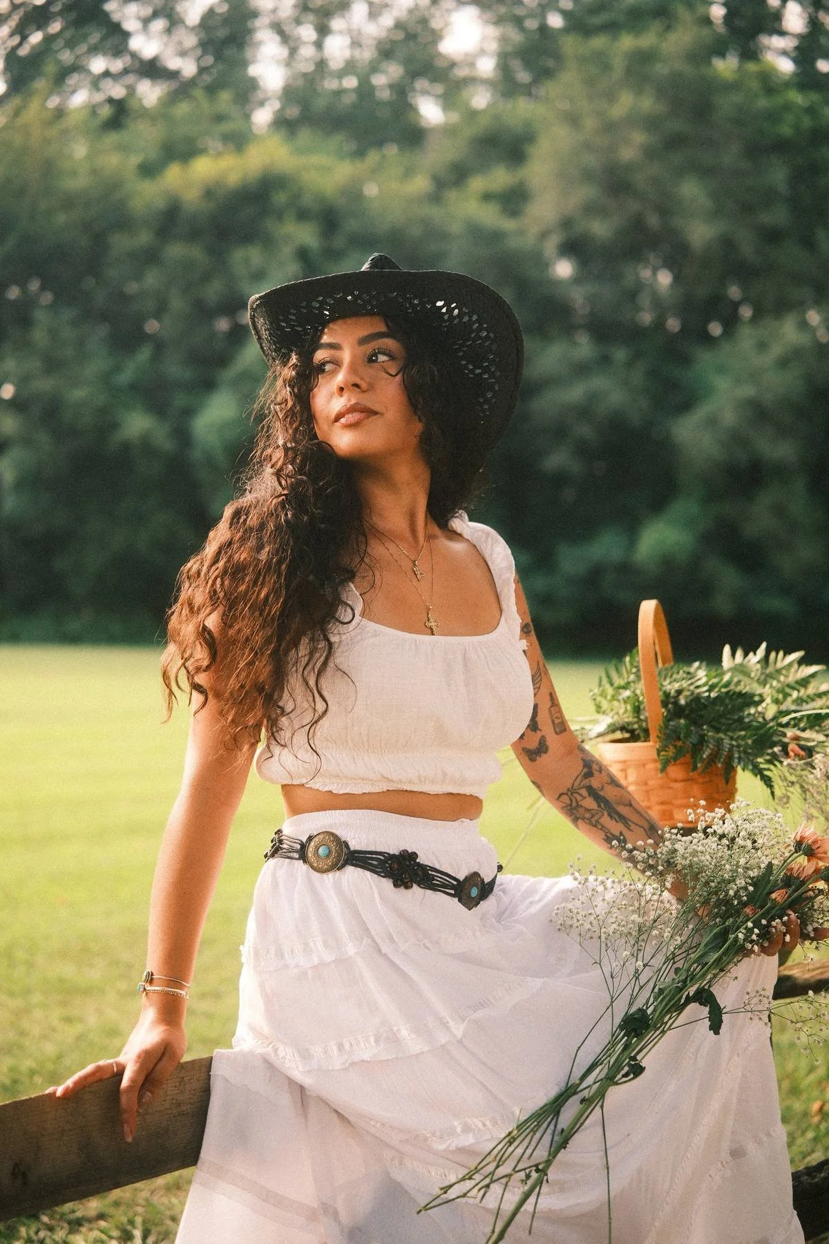 A woman with long curly hair wearing a black wide-brimmed hat and a white sleeveless top, sitting outdoors in a lush green park or garden with trees in the background, surrounded by flowers and a basket of greenery.