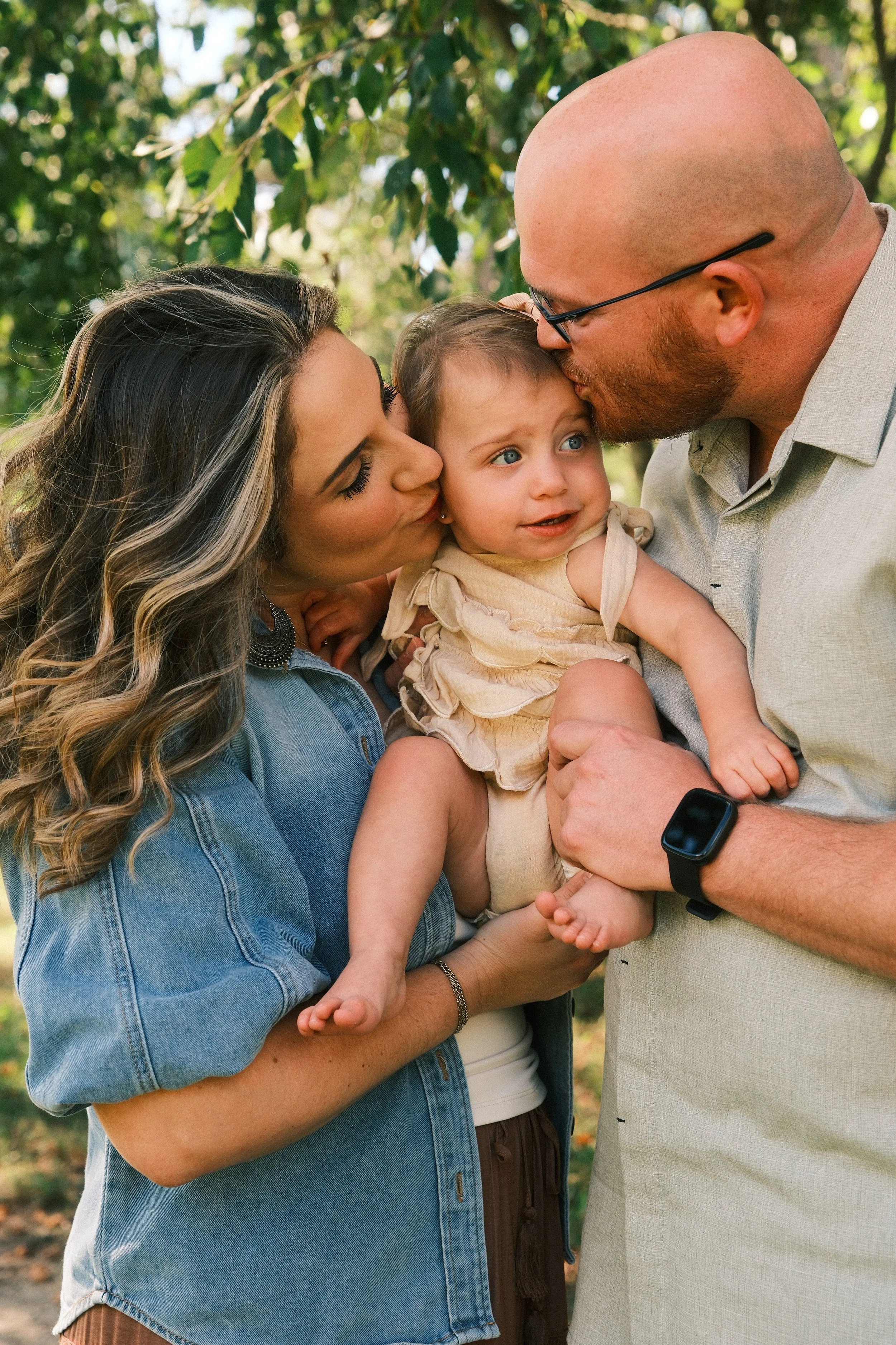 A family of three outdoors with the mother and father kissing their young daughter on the head.