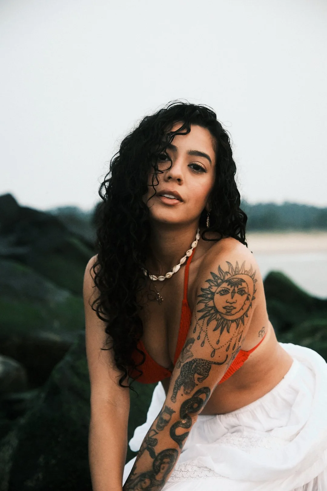 A woman with long curly black hair, tan skin, and tattoos on her right arm, sitting outdoors near rocks with a blurred background of water and sky, wearing an orange top, white skirt, a shell necklace, and earrings.