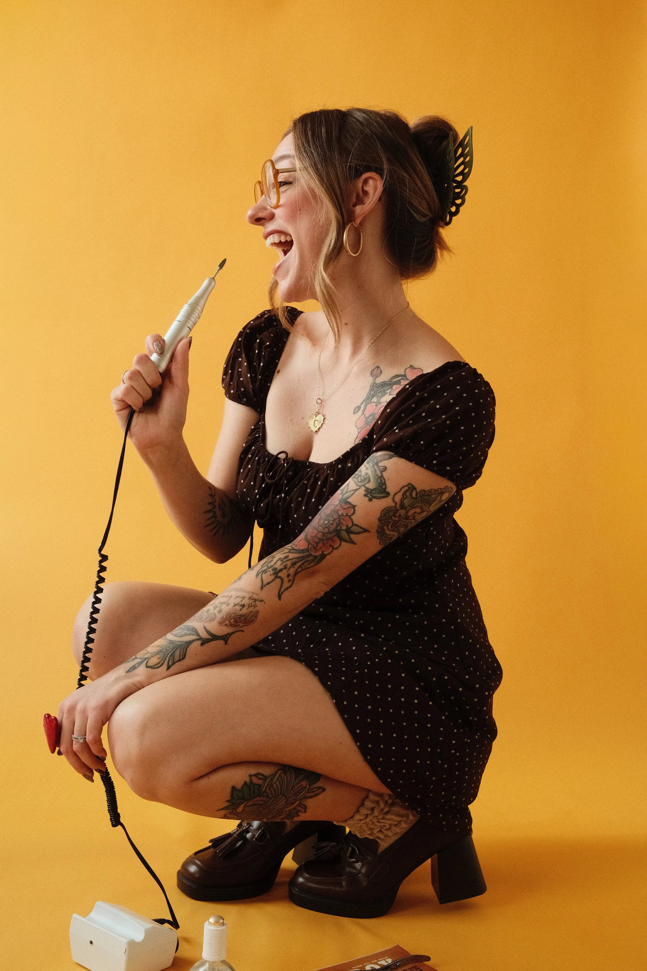 A woman dressed in a black polka dot dress, wearing glasses and earrings, with tattoos on her arms, sitting on a stool against a yellow background, smiling and holding a dental tool near her open mouth.