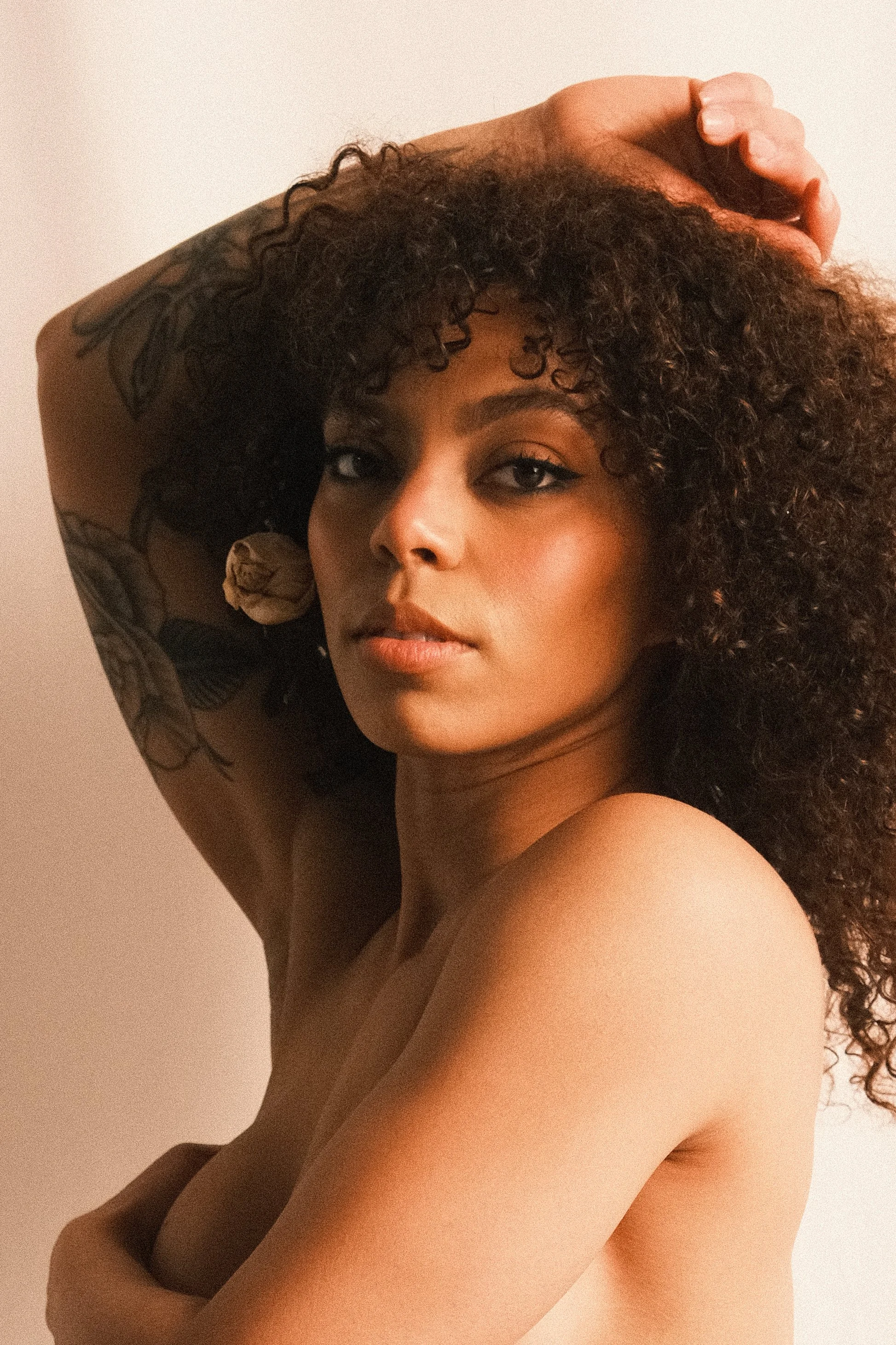 A woman with curly hair and tattoos on her arm posing with her arm raised behind her head against a plain background.