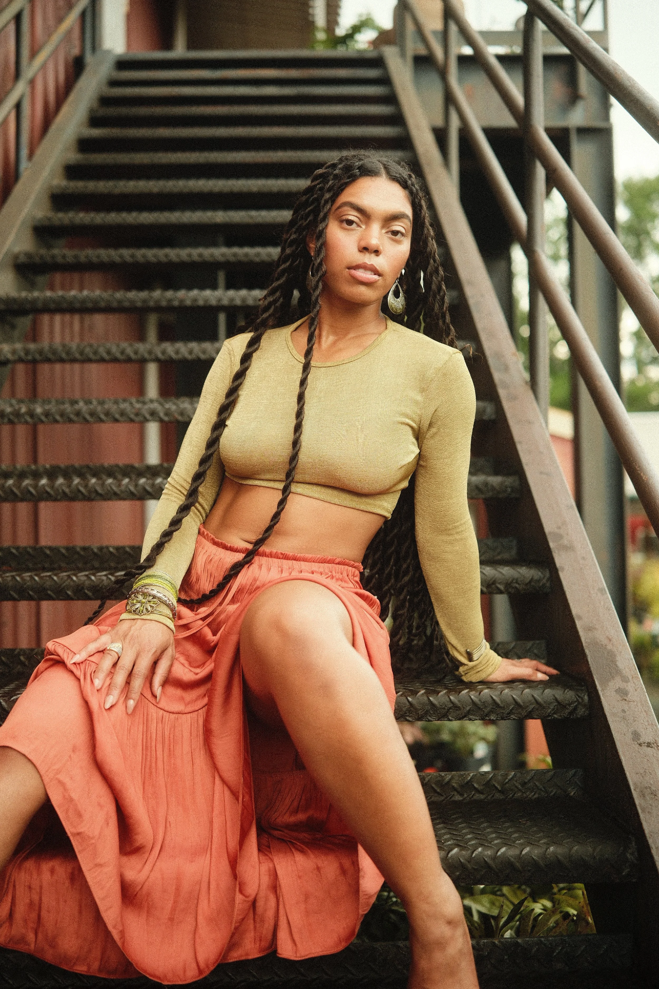 A woman with long braided hair wearing a yellow long-sleeve crop top and orange pants, sitting on a metal staircase outside.