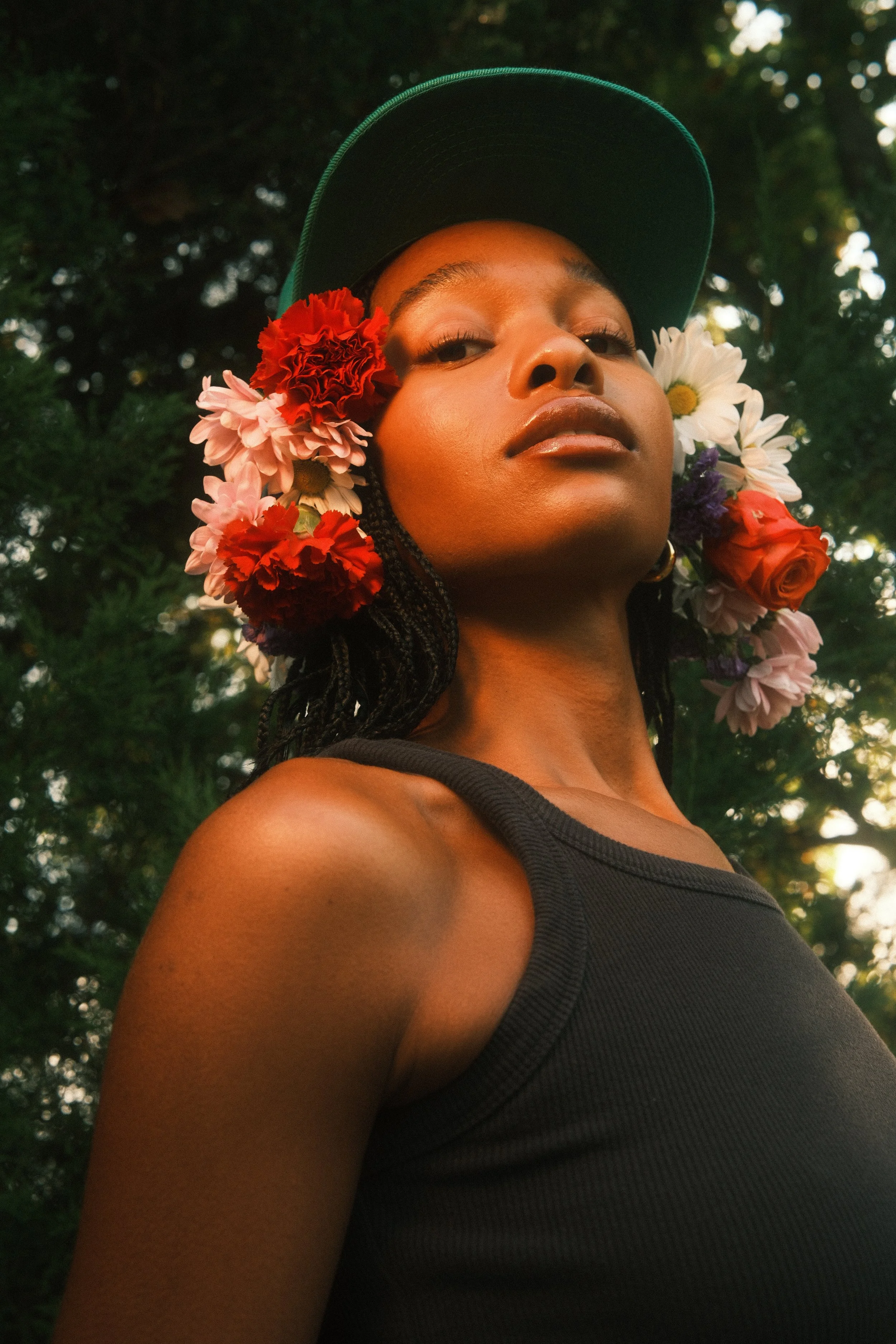 Close-up of a woman wearing a dark green cap and a black tank top, with flowers in her hair and a background of green trees.