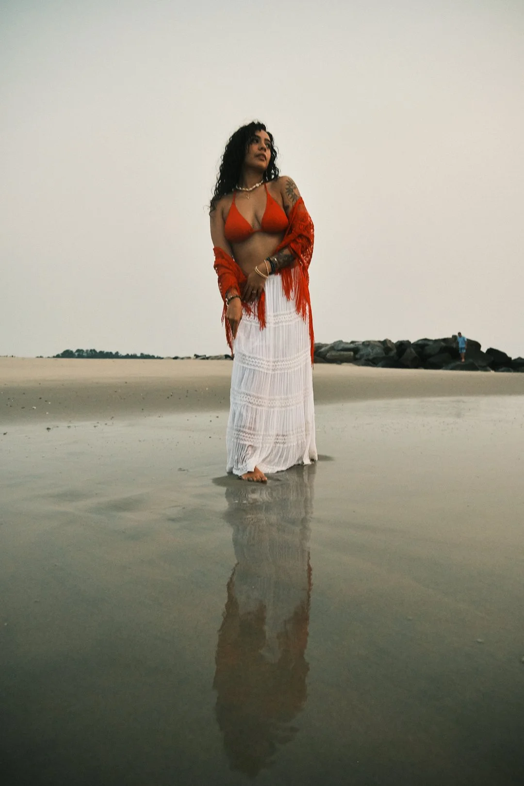Woman standing in shallow water at the beach, dressed in a red bikini top, white maxi skirt, and red shawl, with clouds in the sky and rocks in the background.