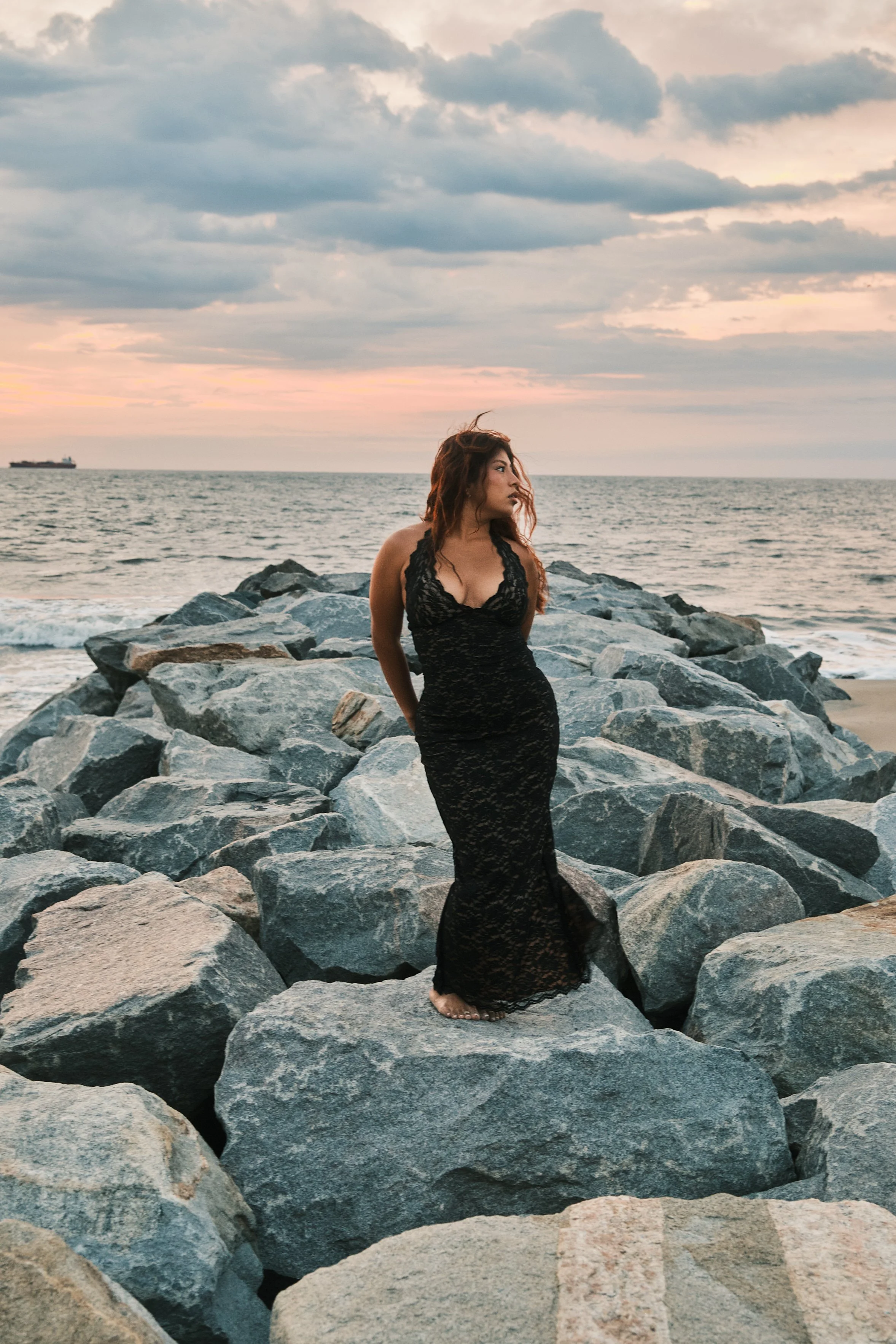 A woman in a long black lace dress standing on large rocks by the ocean during sunset, with cloudy sky and a ship in the distance.