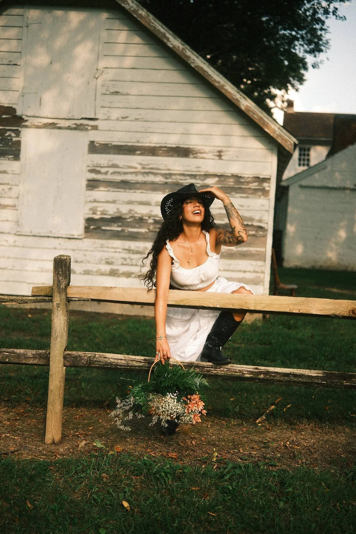A woman dressed in a white dress and black cowboy boots sits on a wooden fence in front of a weathered, white wooden building. She wears a large black hat and is smiling with one hand on her hat. She holds a basket of flowers and greenery, and has ta