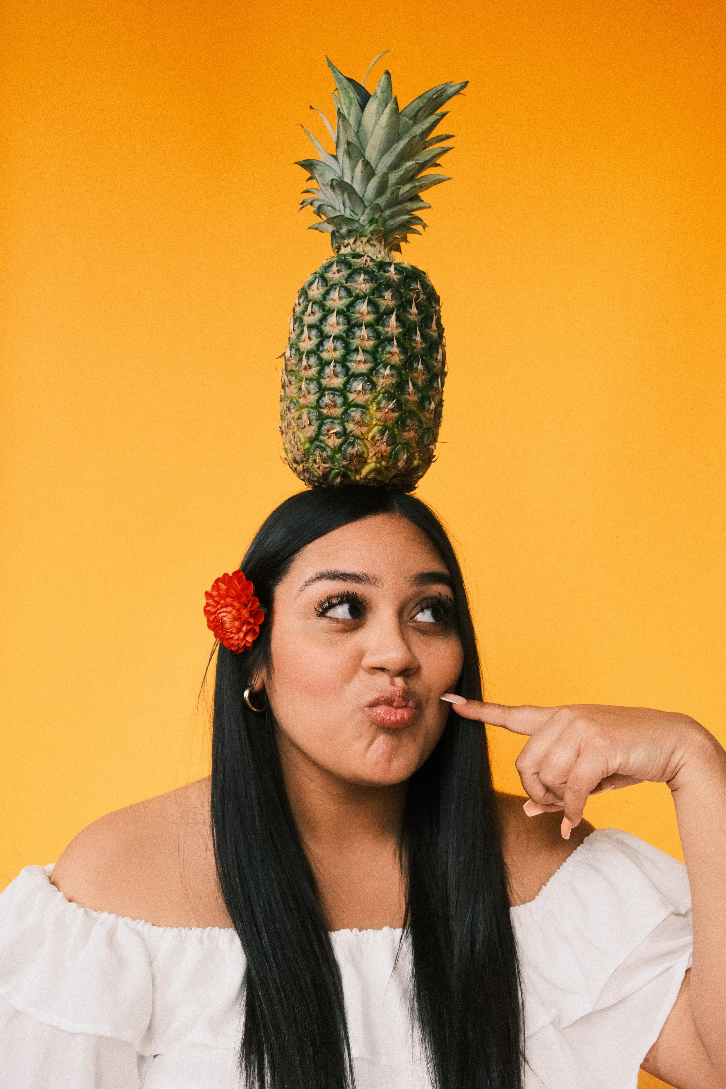 Young woman with long black hair, wearing a white off-shoulder top and a red flower in her hair, balancing a pineapple on her head against a yellow background, pointing at her cheek with a playful expression.