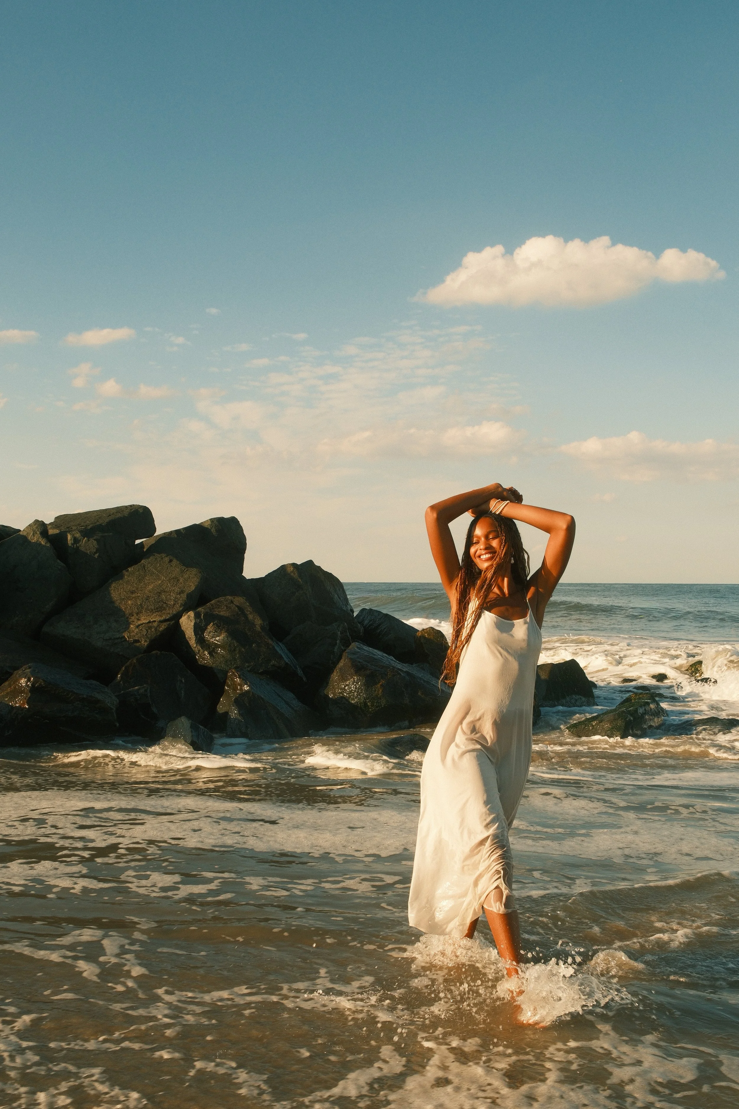 Woman with long hair and a white dress standing in ocean water near large rocks, smiling with arms raised above her head, under blue sky with clouds.