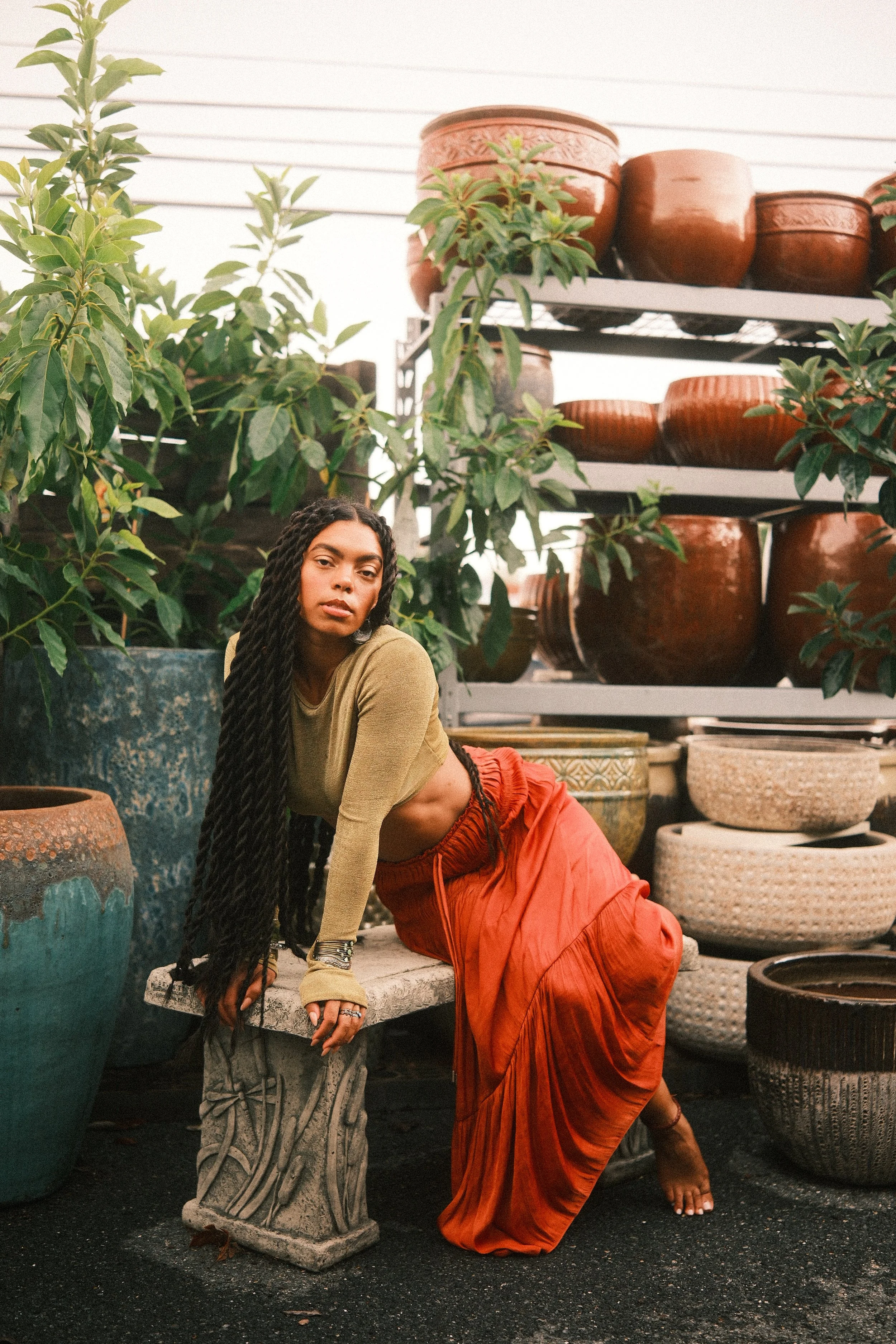 A woman with dark hair styled in long twists, wearing a green crop top and orange skirt, sitting on a concrete surface outdoors among potted plants and shelves with various large brown ceramic pots.