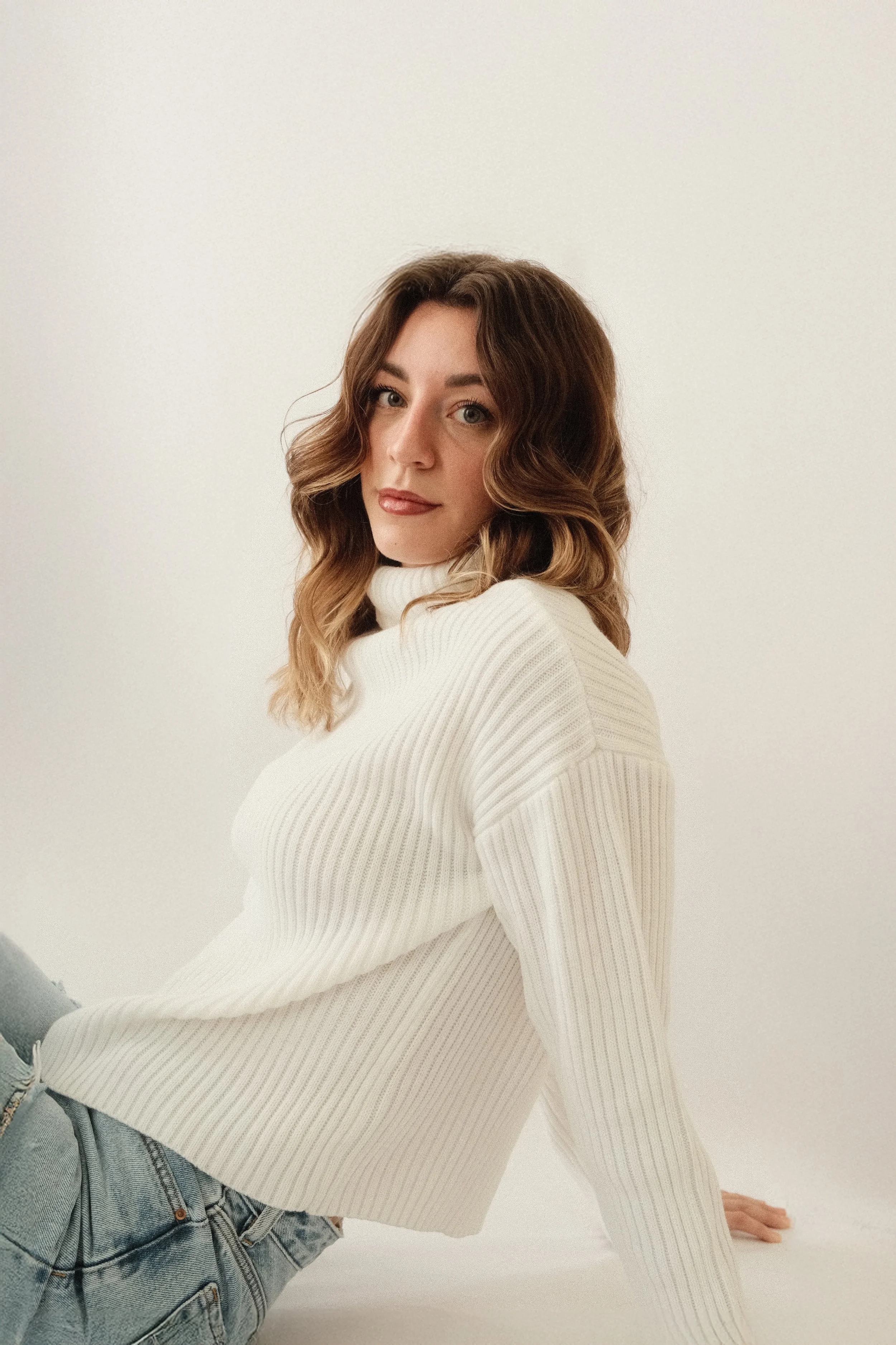 A woman with wavy brown hair wearing a white ribbed turtleneck sweater and light blue jeans.