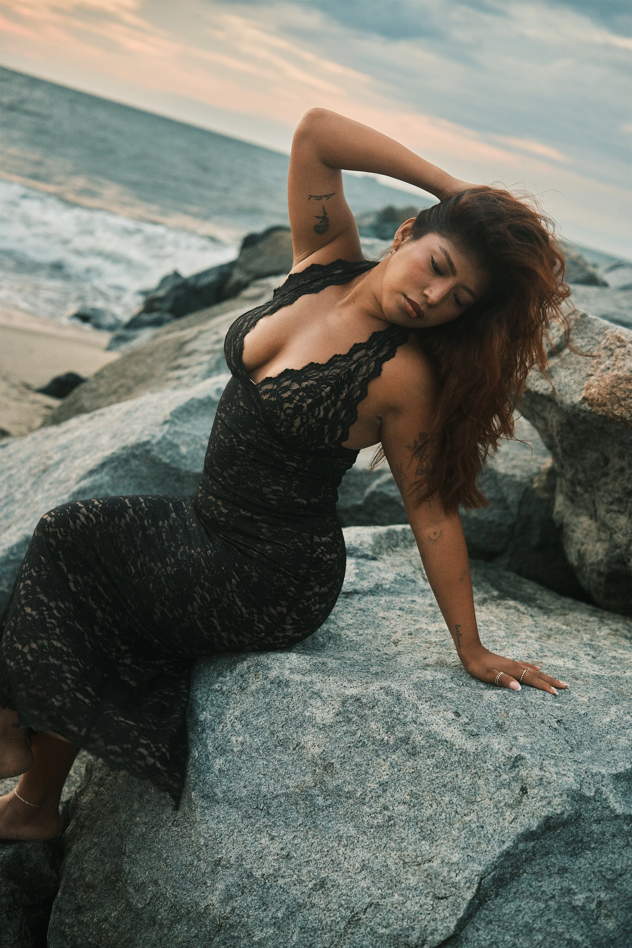 A woman with long curly hair poses on a large rock at the beach, wearing a black lace dress, with the ocean and a sunset sky in the background.