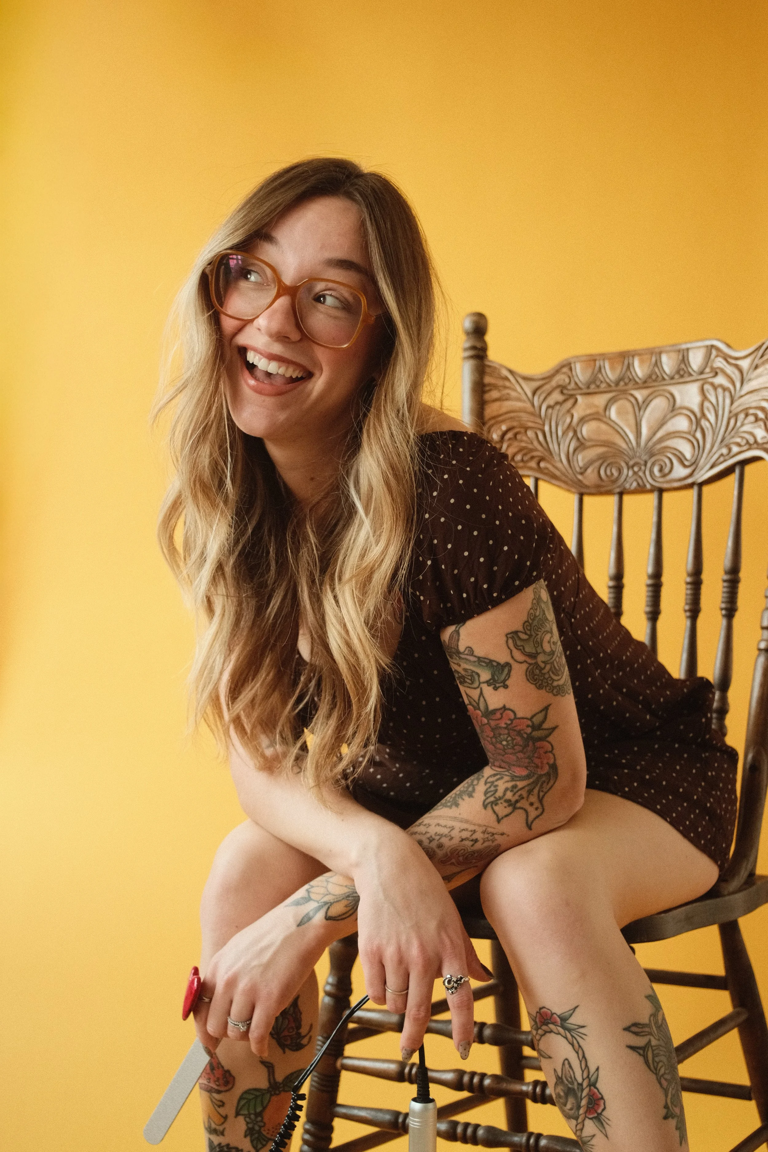 A woman with long wavy hair, glasses, and tattoos on her arms and legs, sitting on a wooden chair against a yellow background.