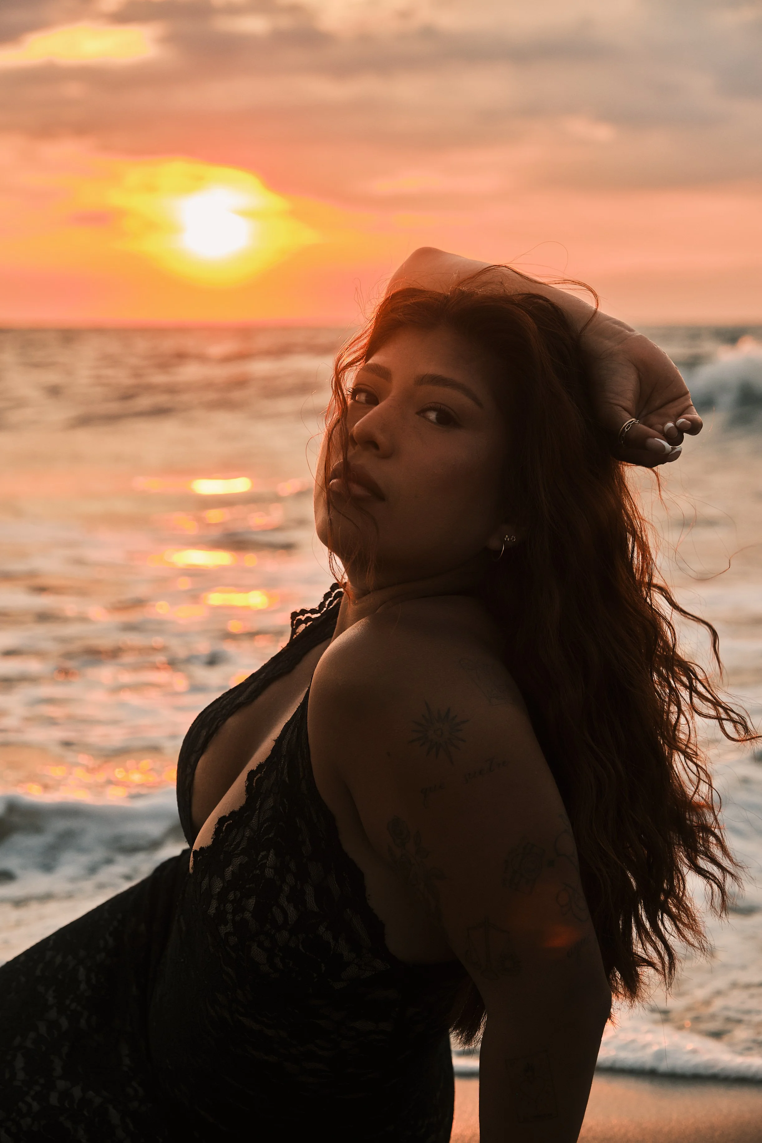 A woman with long hair and tattoos on her arm posing on the beach at sunset, with the ocean and colorful sky in the background.